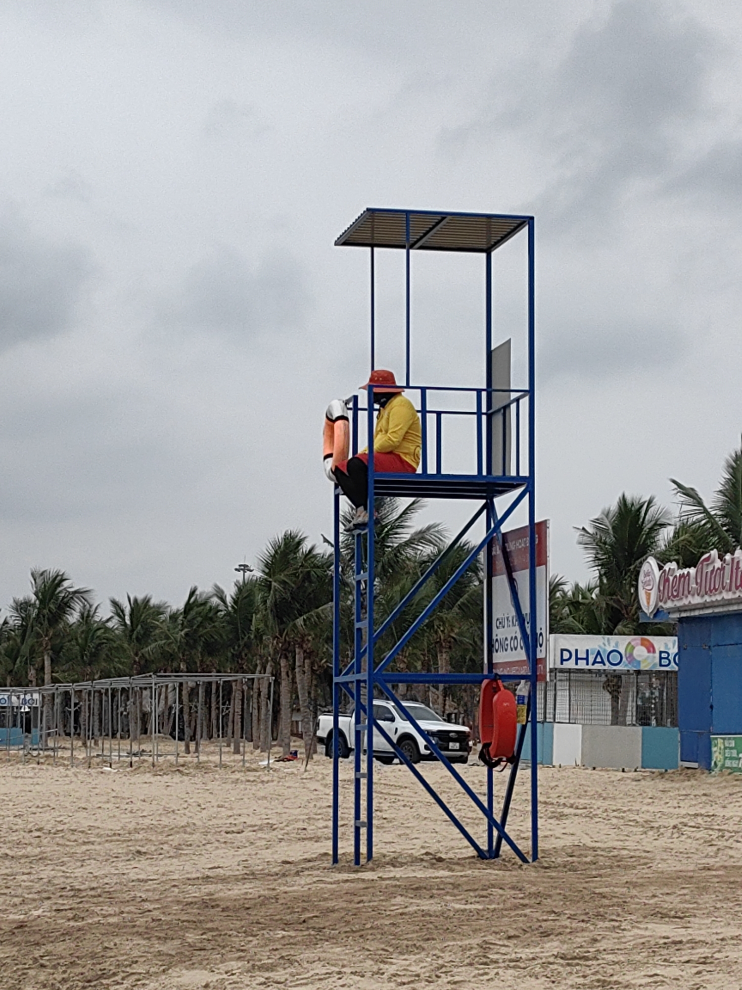A lifeguard sits on a blue metal lifeguard tower on a sandy beach under an overcast sky. The lifeguard wears a red hat, a yellow shirt, and black pants, with a white and orange life preserver next to them. Palm trees line the background. To the right of the tower is a blue building with signage in Vietnamese, "KEM TƯƠI Ý" and "PHAO BOI". Behind the tower is a white pickup truck. The beach is in Bai Chay Ward, Vietnam.