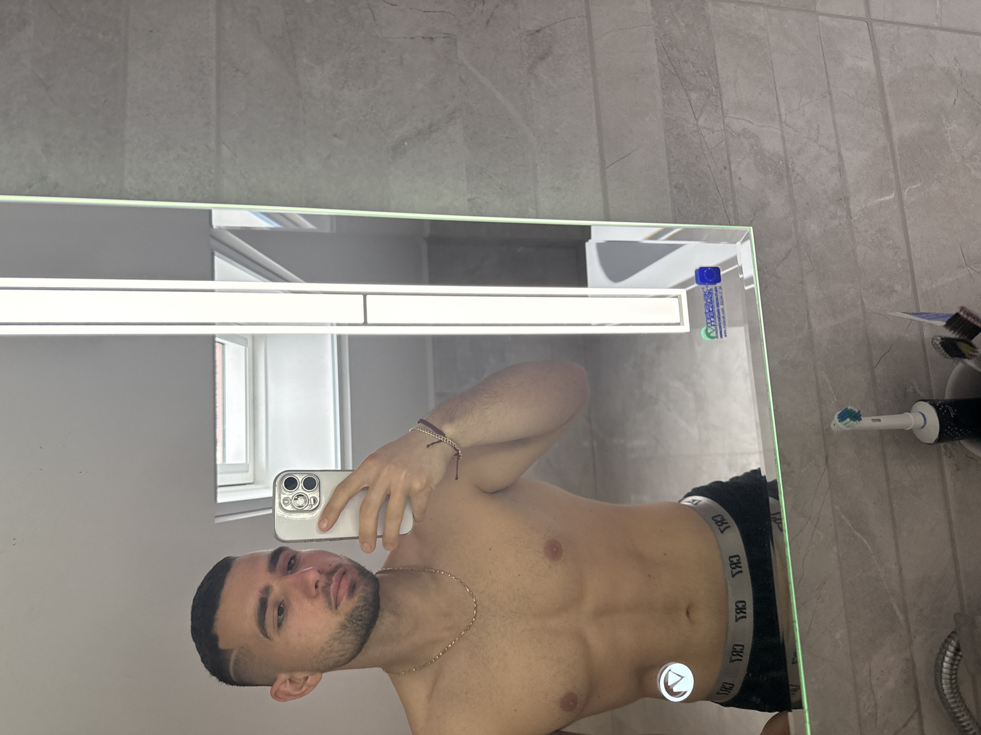 A shirtless man is taking a mirror selfie in a bathroom with gray marble-like tiles. He is wearing dark-colored underwear with a light-colored waistband. He is holding a white smartphone with multiple cameras in his left hand. The bathroom contains a lighted mirror with an electric toothbrush and toothpaste on the right side of the mirror. The man has a gold chain necklace and a bracelet on his left wrist. The location is a bathroom in Gabrovo, Bulgaria.