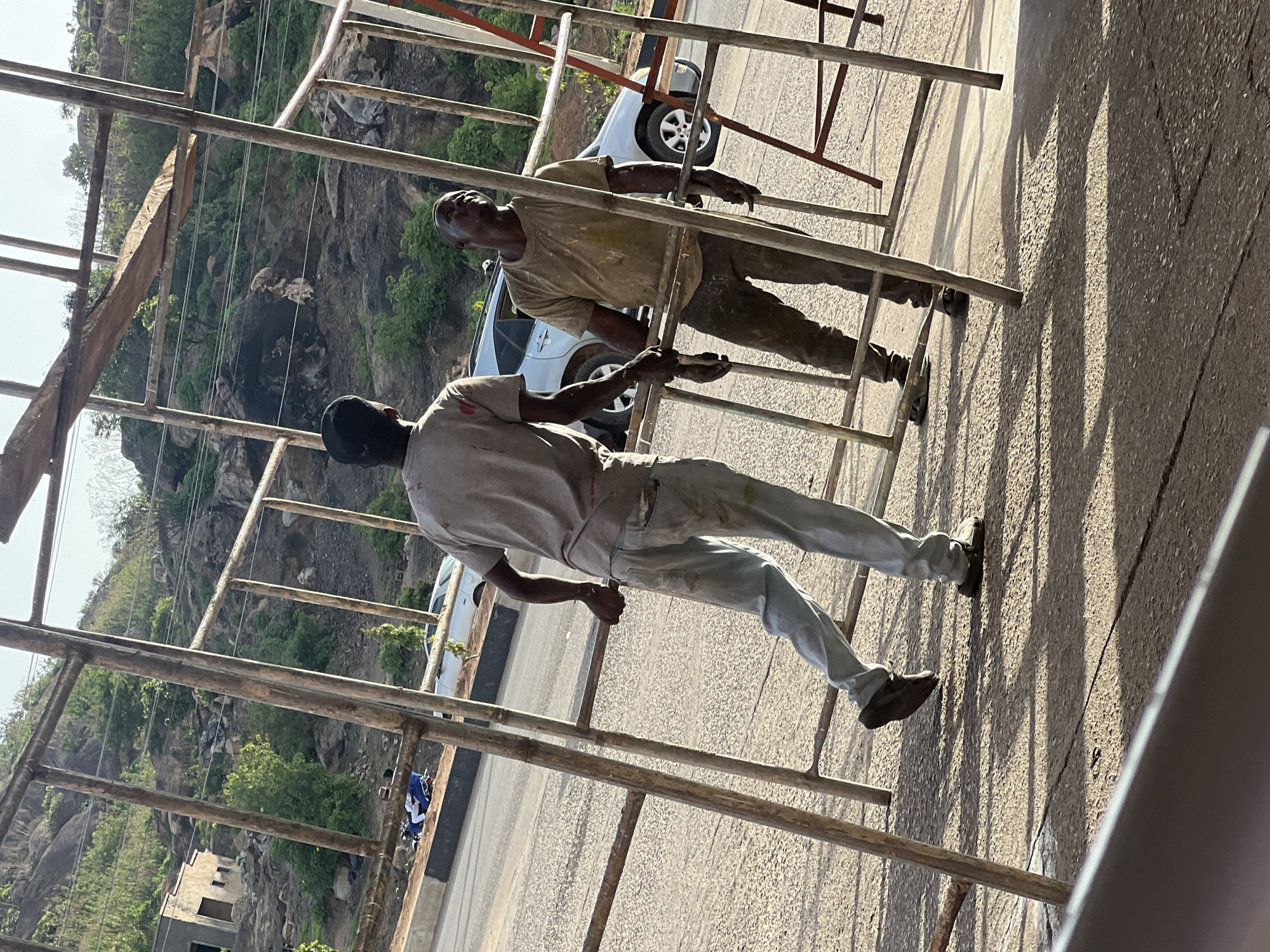 Two men are working on scaffolding. The man closer to the viewer wears a light-colored shirt and pants. The man further away wears darker clothing. A car is parked in the background. The location is Gura Top, Nigeria, based on the image's origin. The environment includes a roadside with hills and vegetation in the distance.