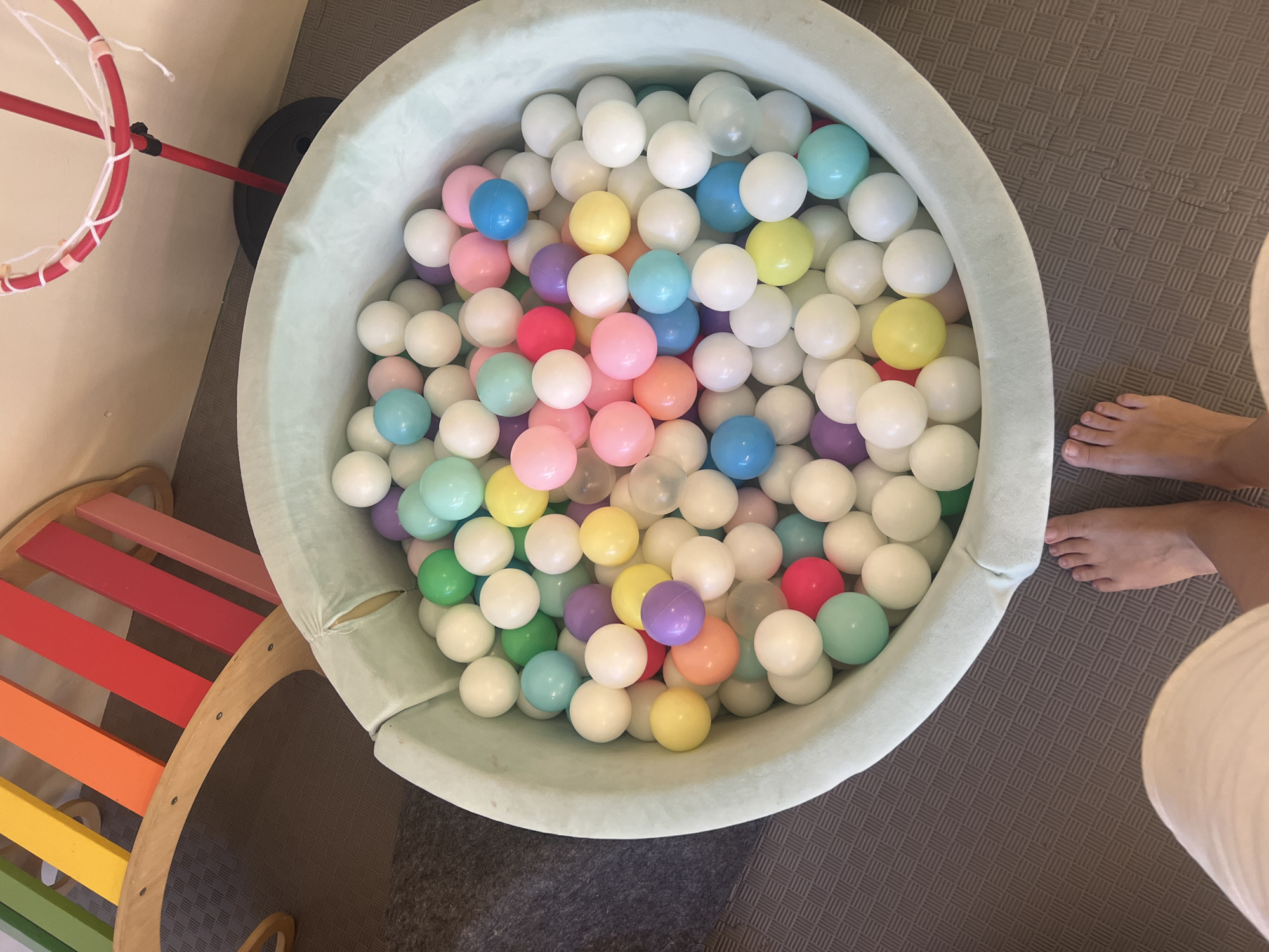 A high-angle, full shot captures a light green ball pit filled with numerous plastic balls of various colors, including white, light blue, green, yellow, pink, purple, and orange. The ball pit is situated on a grey, textured mat. To the left of the ball pit, a wooden rainbow climber with colorful planks is partially visible. To the upper right, a red toy basketball hoop with a white net stands behind the ball pit. In the foreground, the lower legs and feet of an unseen person are visible, with the left foot positioned to the left of the ball pit and the right foot to the right. The overall scene suggests a child's play area. There is no indication of the location being in Ko Samui District, Thailand.