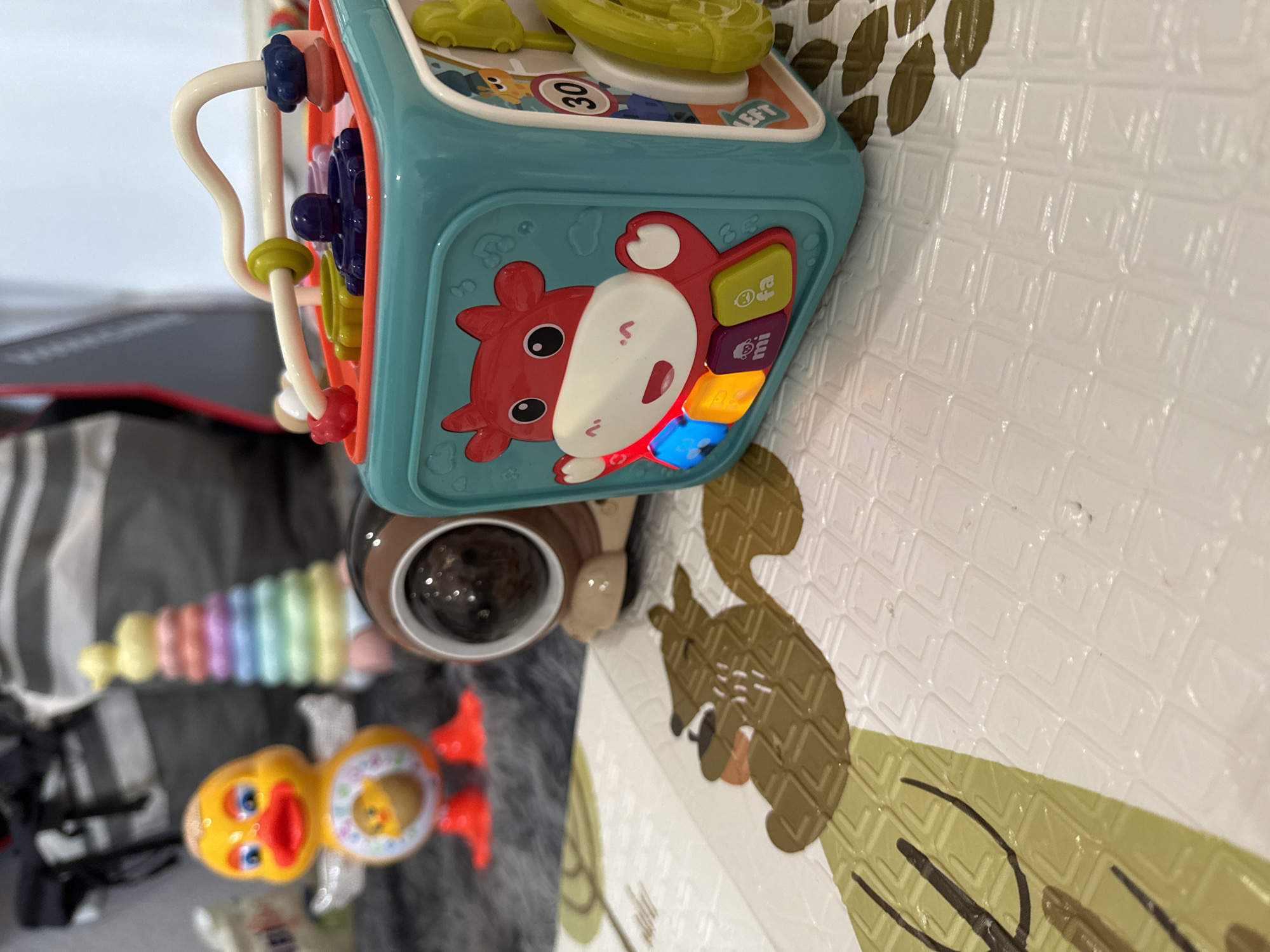 The image shows a close-up of children's toys on a textured play mat. The primary object is a light blue activity cube with a red hippo design on one side and buttons that illuminate in various colors. Other toys include a brown ball with a dark circle on top, a stack of pastel-colored rings, and a yellow duck toy with red feet. The play mat has a white background with a textured pattern and a design of a brown squirrel and green leaves.
There is no information in this image that references Yakutsk, Russia.