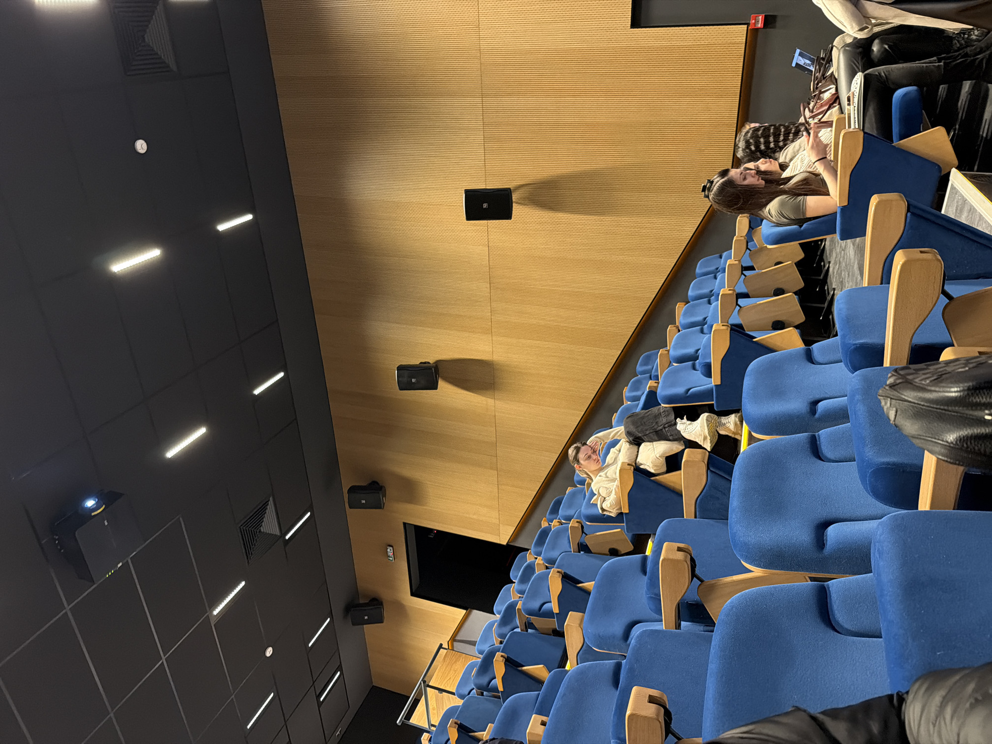This is a photograph of an empty auditorium with blue upholstered seats and wooden armrests. Two young women are seated in the back row, both looking at their phones. The walls are paneled with light wood, and there are speakers mounted on the walls. The ceiling is dark with recessed lighting, ventilation grates and a projector. In the bottom left corner is a wall mounted projector. The photograph was taken at the Dom na Kinoto (House of Cinema) in Sofia, Bulgaria.