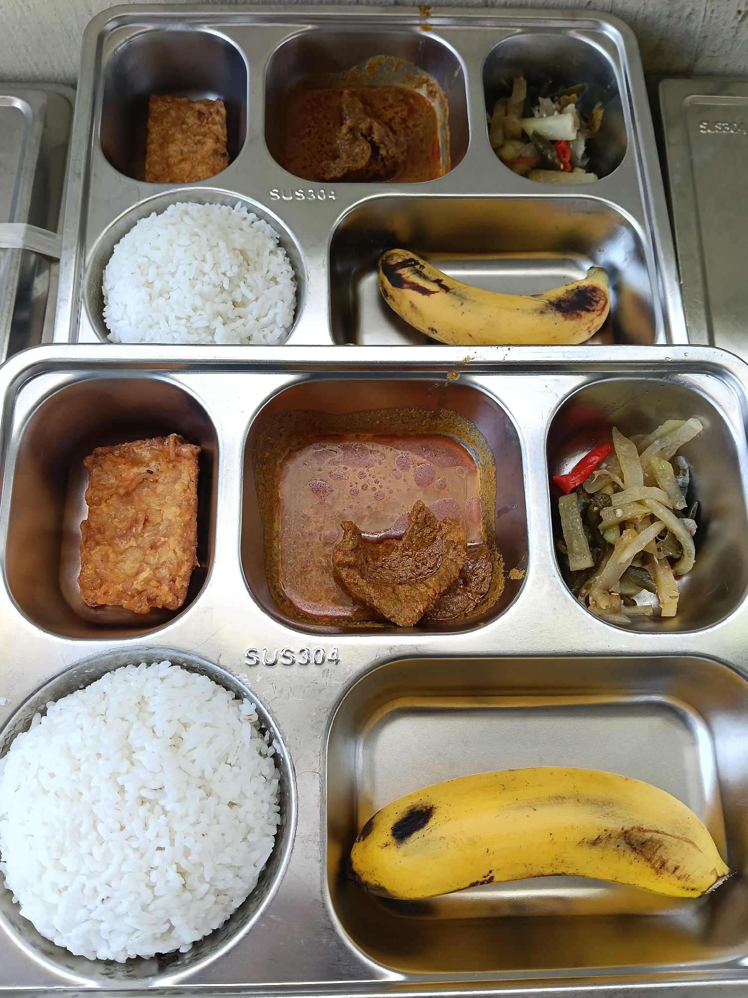 The image shows two stainless steel food trays, each with five compartments, stacked on top of each other. Each tray contains a meal consisting of rice, a square piece of fried tempeh, a serving of beef rendang, a portion of pickled vegetables, and a banana. The text "SUS304" is imprinted on the trays. The image was likely taken in Lhokseumawe, Indonesia, based on the typical cuisine served.