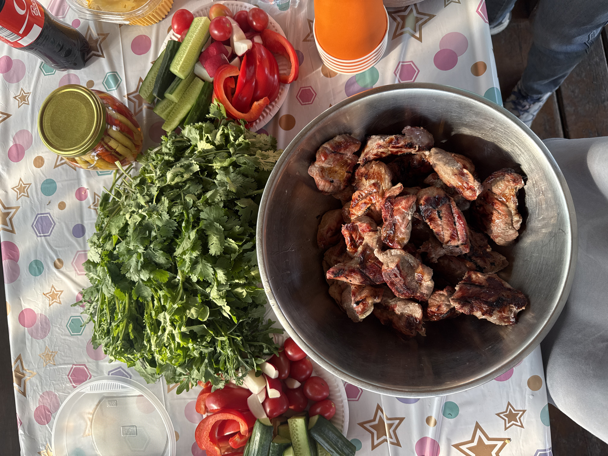 The image shows a spread of food on a table with a patterned tablecloth. A large metal bowl is filled with grilled meat, possibly shashlik. There is a large bunch of cilantro next to the bowl. Plates of sliced vegetables, including cucumbers, tomatoes, radishes, and red bell peppers, are also on the table. A jar of pickled vegetables, an orange cup, and a bottle of Coca-Cola are visible. The scene appears to be a gathering or picnic of some sort. The location information indicates that this image was likely taken in or near the city of Кузнецово, Russia. A person is partially visible in the upper right of the image. The setting is outdoors, as indicated by the lighting.