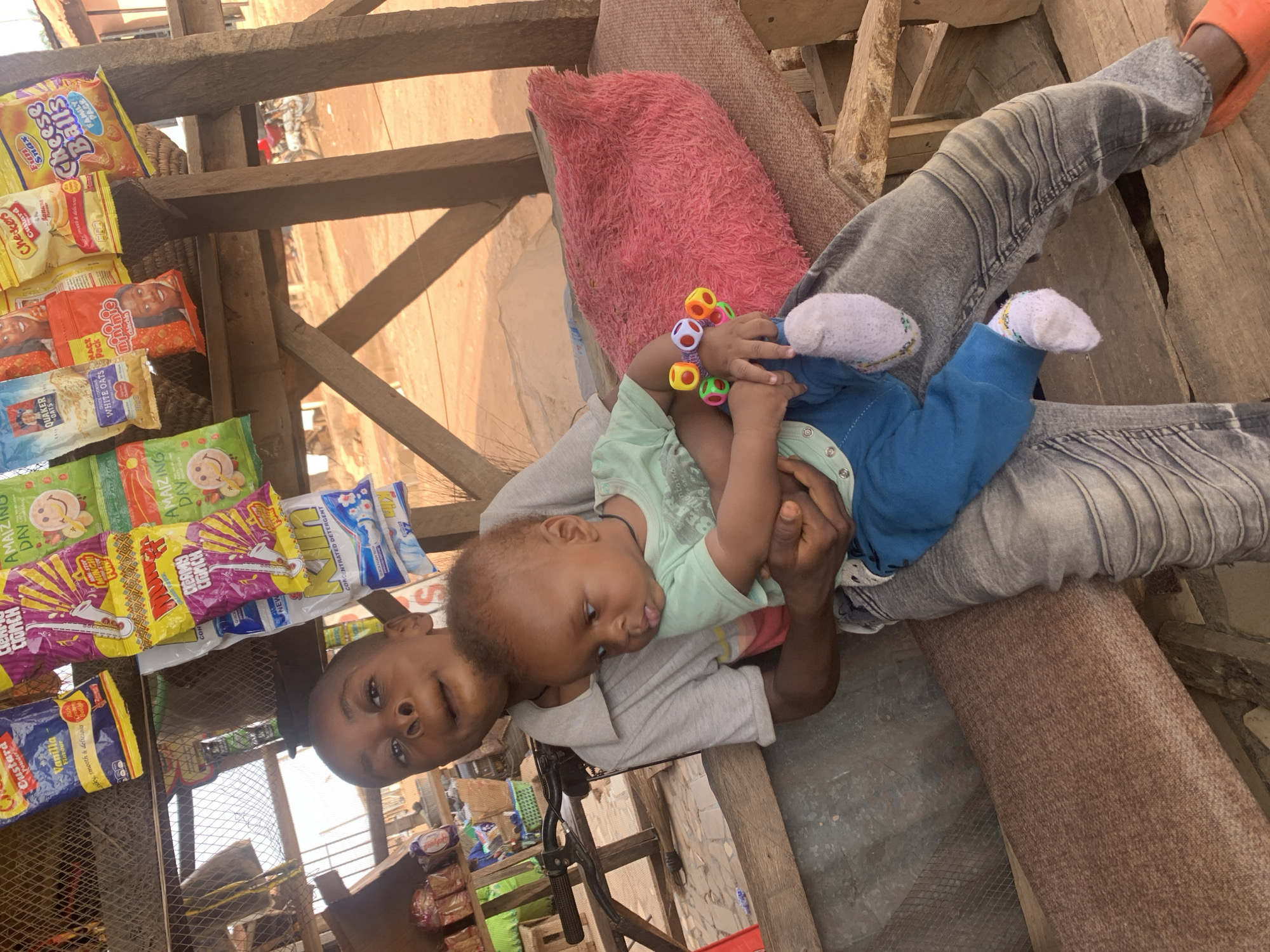 This portrait-oriented image, rotated 90 degrees clockwise, depicts a young person holding an infant within an outdoor market stall or small shop, likely in Sabon Tasha, Nigeria. The central figure is a young individual with dark skin and short dark hair, wearing a light grey t-shirt and faded grey jeans, with orange slip-on shoes. They are seated on a wooden structure, partially covered by a pink furry blanket or pillow, and gaze directly at the camera with a neutral expression.
In their arms, an infant with dark skin, dressed in a light green top, blue pants, and white socks, is held securely. The infant is holding a brightly colored rattle with multi-faceted spherical beads and is looking to the side. Natural daylight illuminates the scene, indicating it is daytime.
The setting is characterized by a makeshift wooden structure, possibly a market stall, with shelves and hanging racks displaying various packaged goods. Visible products include "Cheese Balls," "Checkers Custard," "minimie chinchin" snacks, "Quaker Oats," "AMAIZING DAY" snacks, "CREAMY CORNCH" snacks, and "Kin CONCENTRATED DETERGENT." These items are arranged for sale, suggesting a commercial environment. In the blurred background, glimpses of other structures and a motorcycle further contextualize the outdoor, bustling market atmosphere. The scene captures a moment of daily life, possibly work or rest, in a local retail space.
