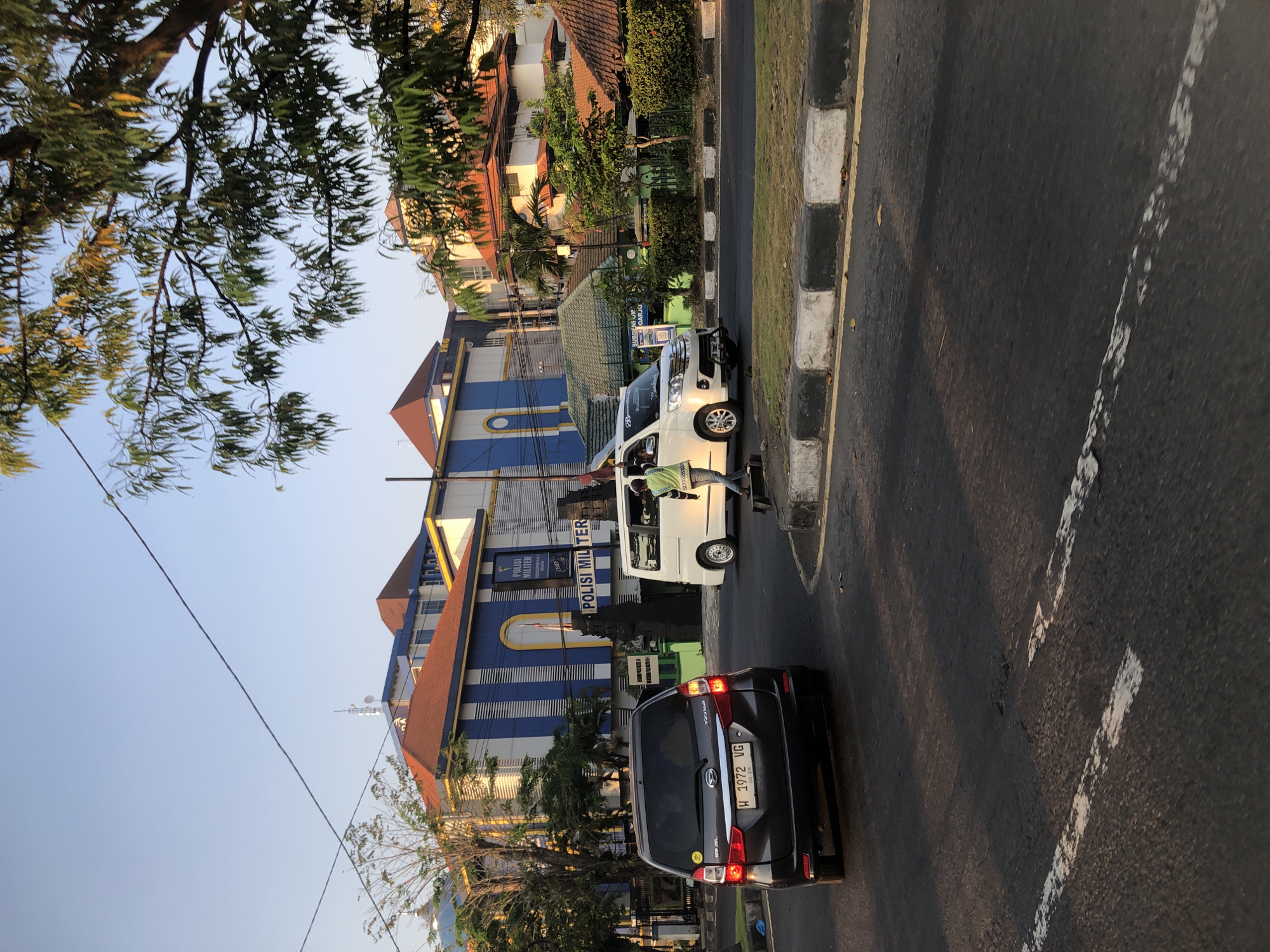 This image captures a street scene in the city of Larangan, Indonesia, likely during the late afternoon due to the warm lighting. In the foreground, a dark gray Daihatsu Xenia is parked on the left side of the asphalt road. The car has a license plate that reads "W 1972 VG". Further down the road, a white van is in motion. A person wearing a bright green vest over a dark shirt and pants is visible near the van, appearing to be standing on a skateboard or scooter.
The background features a large, blue and white striped building with an orange roof, identified by signage as "POLISI MILITER", indicating it is a military police station. The building has a large arched entrance with a yellow outline. To the right of this building, there are smaller structures with orange-tiled roofs and green foliage surrounding them. Trees with green leaves, some yellowed, line the side of the road and the buildings, with branches extending into the sky. Power lines are visible crisscrossing the frame. The road is marked with white dashed lines and has a curb with black and white painted borders. The overall environment suggests a moderately busy urban area with governmental or official buildings.