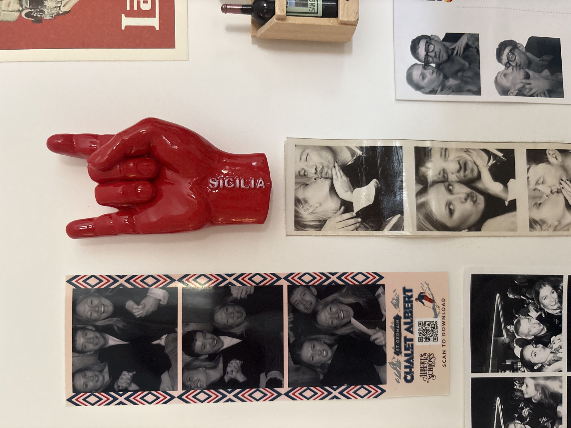 A close-up shot of a white wall adorned with various items, likely a collection of personal mementos. From the top-left corner, a partial view of a red and white poster is visible. Adjacent to it is a wooden miniature wine rack holding a small bottle of Sangiovese wine. A black-and-white photo strip captures a couple in a playful pose, kissing. A red, ceramic hand-shaped decoration with the word "SICILIA" imprinted on it is located on the left side of the frame. Below it, another black-and-white photo strip depicts a group of three people. Next to it, a souvenir strip from the Chalet Albert St-Germain ski resort, including a QR code. Another black and white photo strip is next to the resort strip.
The items are affixed to a white wall. Based on the name "Albert" on the souvenir strip, and the name "St-Germain", it can be speculated that the location may be the Chalet Albert, a Swiss-themed restaurant located in London, United Kingdom.