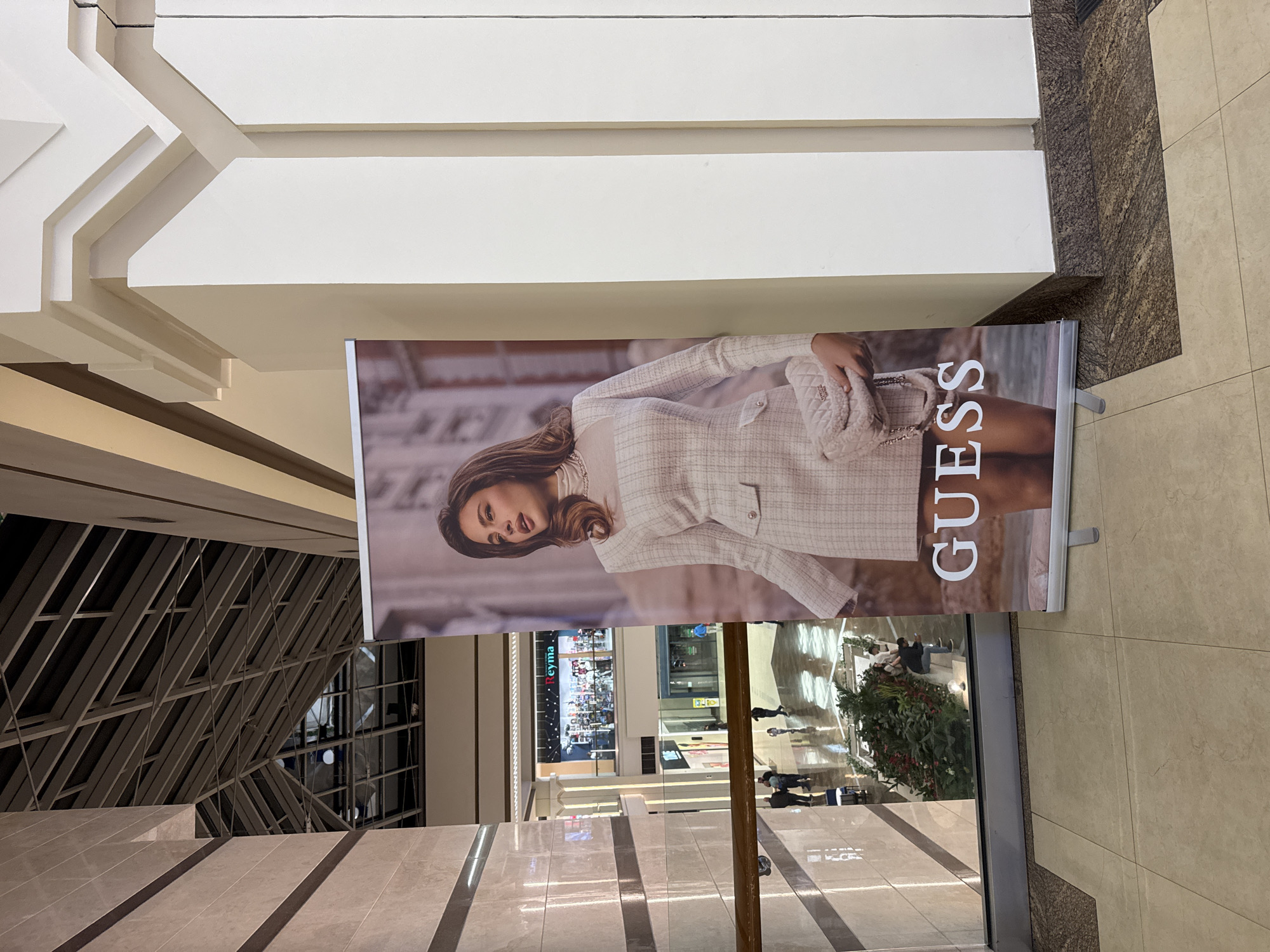 This image shows a Guess advertisement banner in a Yerevan, Armenia shopping mall. The advertisement features a woman in a tweed jacket, holding a handbag, with the Guess logo vertically oriented next to her. The banner is placed along a walkway with a partial view of other shops and mall traffic. Overhead, a decorative ceiling structure is visible. The floor is tiled, and a structural element is on the left with similar tiling.