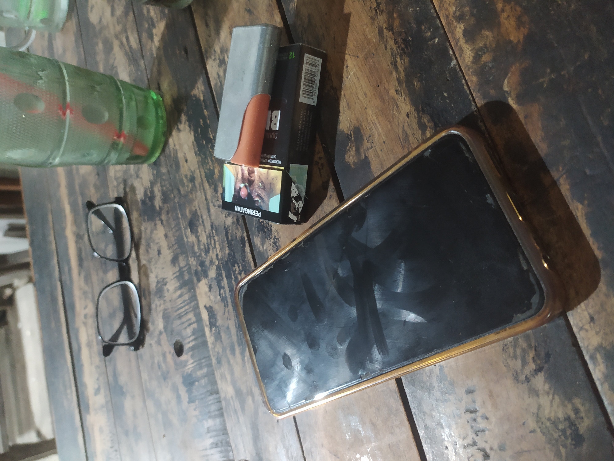 A smartphone, a pack of cigarettes with a metal case, a pair of eyeglasses, and a green glass with a red straw are arranged on a weathered wooden table. The scene appears to be indoors, possibly at a roadside cafe in Tuntang, Indonesia. The lighting suggests it is evening or nighttime. The smartphone screen is black and smudged, indicating it is not in use. The cigarette pack has Indonesian text and images of eyes, typical of health warnings. The eyeglasses are black-rimmed and resting on the table. The green glass contains a clear liquid and a red straw. The wooden table has visible grain and wear marks, suggesting it is an outdoor or rustic setting.