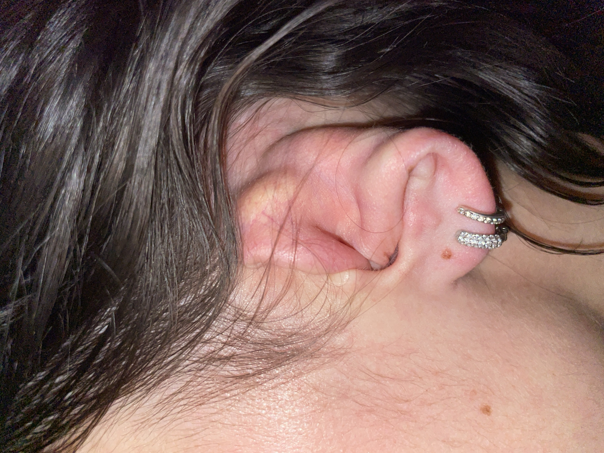 A close-up image depicts a person's ear adorned with a double conch piercing, each ring embellished with small stones. The earlobe and surrounding skin display various veins and capillaries. Short, dark, wet, wavy hair frames the ear. A mole is visible on the neck near the base of the ear. The image does not provide any information about the location in the city of Izhevsk, Russia.