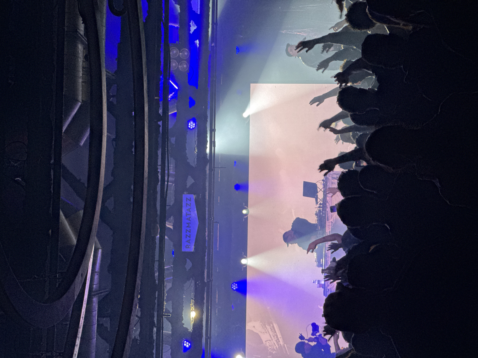 The image captures a concert at Razzmatazz in Barcelona, Spain. A DJ is performing on stage, visible behind a crowd of concertgoers with their hands raised. Stage lighting in hues of white and blue illuminates the scene. The venue's industrial architecture, including exposed beams and metal structures, is visible on the side. A sign displaying the venue's name, "RAZZMATAZZ," is present.