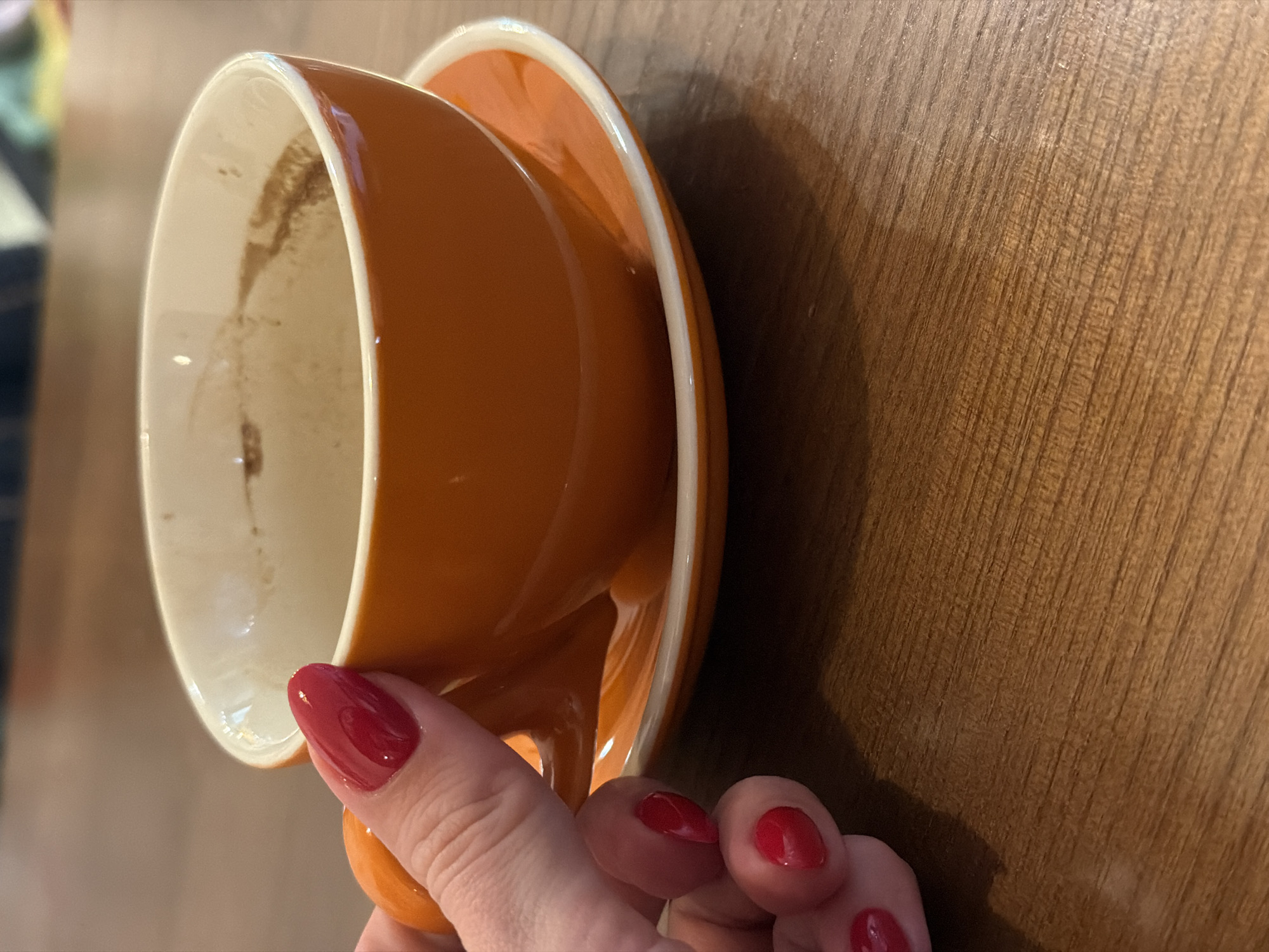 A partial close-up depicts a hand holding an empty, orange ceramic coffee cup and saucer. Residue is visible inside the cup. The hand is fair-skinned with red polished nails. The cup and saucer are positioned against a textured brown wood surface. The location is the city of Dnipro, Ukraine.