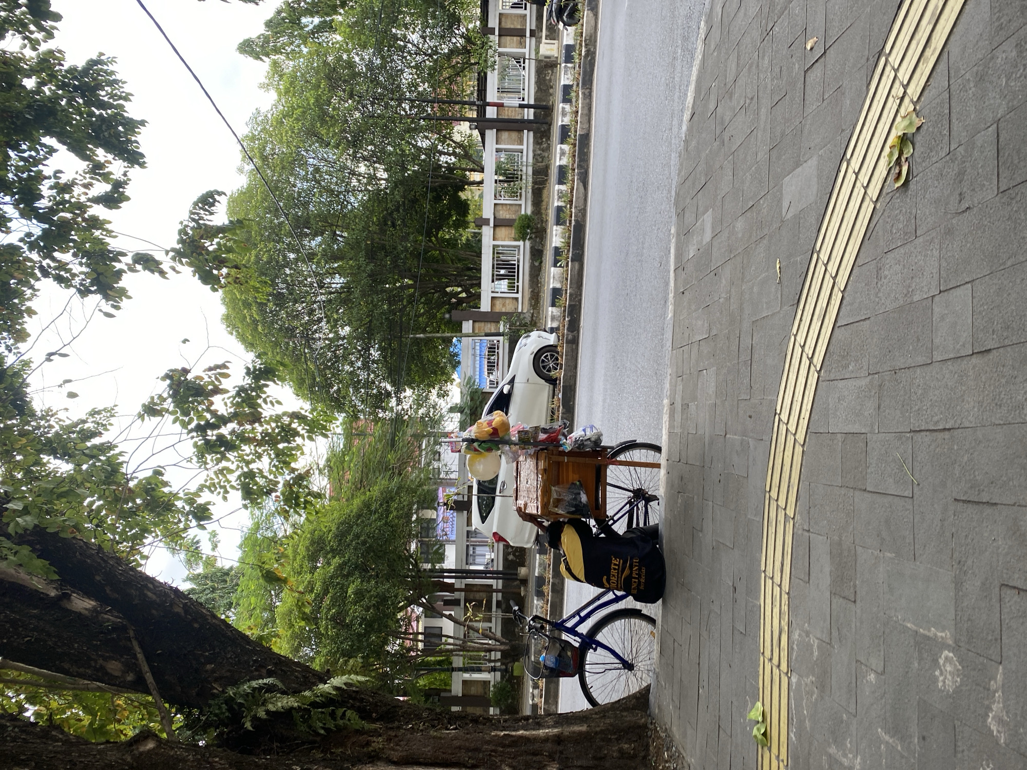 In Banjarbaru, Indonesia, a street scene unfolds with a focus on a blue bicycle with a wooden cart attached. The bicycle is parked on a sidewalk paved with gray tiles and a distinctive yellow tactile paving strip. A dark backpack with yellow lettering, possibly a brand or slogan, is placed on the ground behind the bicycle. A person, wearing a yellow and black jacket, is crouched next to the cart, appearing to be engaged in some activity related to the cart's contents.
The cart itself is brown and appears to be a mobile vending unit, with various items displayed on top, including what look like balloons and packaged goods. A white car is visible in the background, driving on the road which runs alongside the sidewalk. The road has a black and white striped median. Behind the road, a fence with rectangular openings and a light-colored wall runs along a property. Trees with dense green foliage line the street, some casting shadows. The sky is overcast, indicating a possibly cloudy day. The overall impression is of a typical urban street scene in a tropical climate, with a street vendor preparing for business.