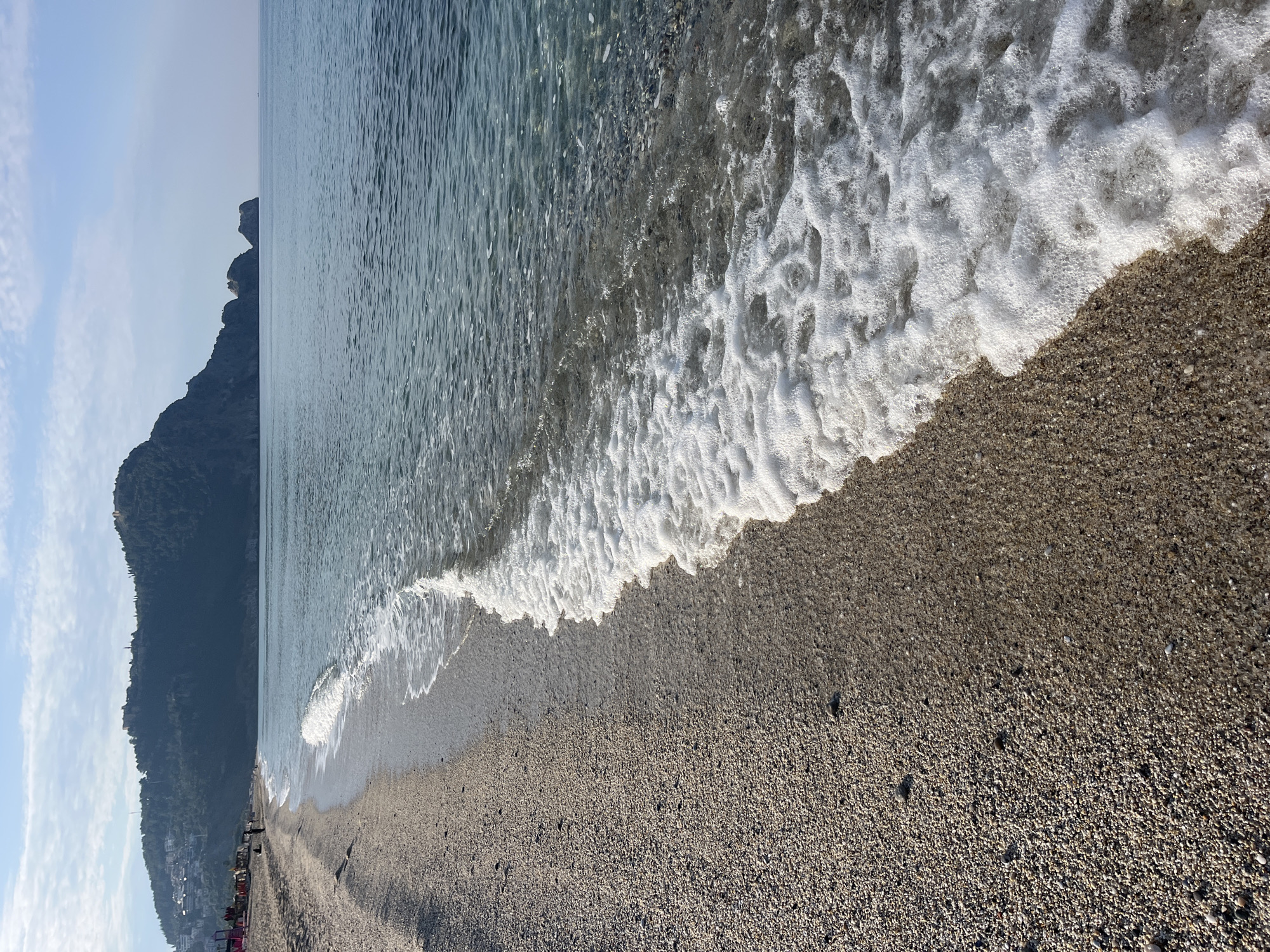 The image shows a beach in Alanya, Turkey. The beach consists of small pebbles. Waves are crashing on the shore, creating white foam. In the background is a large, green mountain range. The sky is pale blue with some clouds. In the distance, beach umbrellas and buildings can be seen.