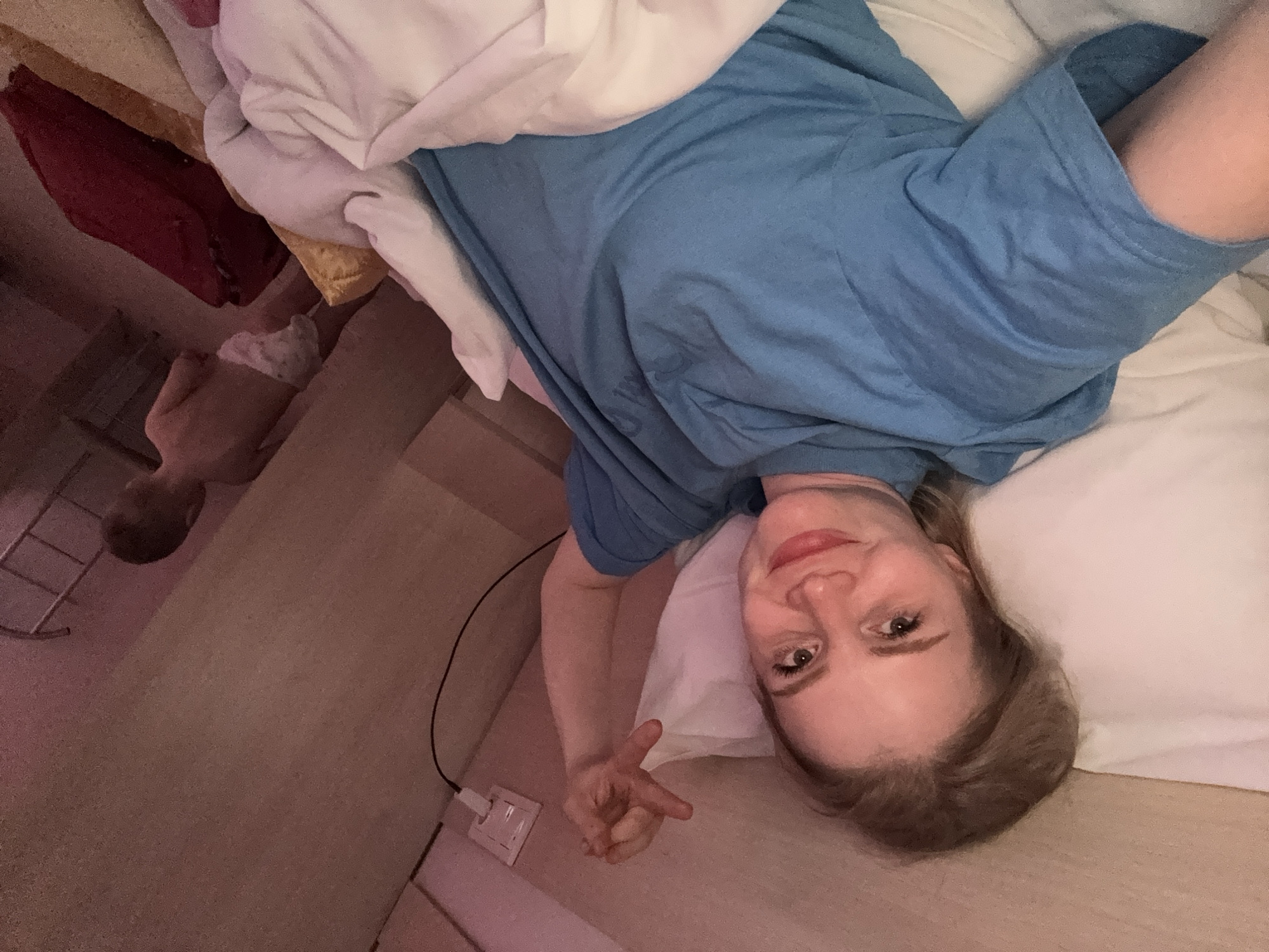 A woman is lying in bed, wearing a blue t-shirt and looking at the camera. Her right arm is raised, and she is making a gesture with her fingers. A child, wearing only a diaper, is visible in the background, standing and facing away from the camera. The room is dimly lit, with pinkish lighting from an unknown source. An electrical outlet with a plug and cord is visible on the wall to the right of the woman. The bed is covered with white and pink bedding, and a red pillow is visible on the right side of the bed. The overall impression is of a casual, domestic scene, possibly in a hotel room or a bedroom. The location in Saint Petersburg, Russia, is not discernible from the image alone.