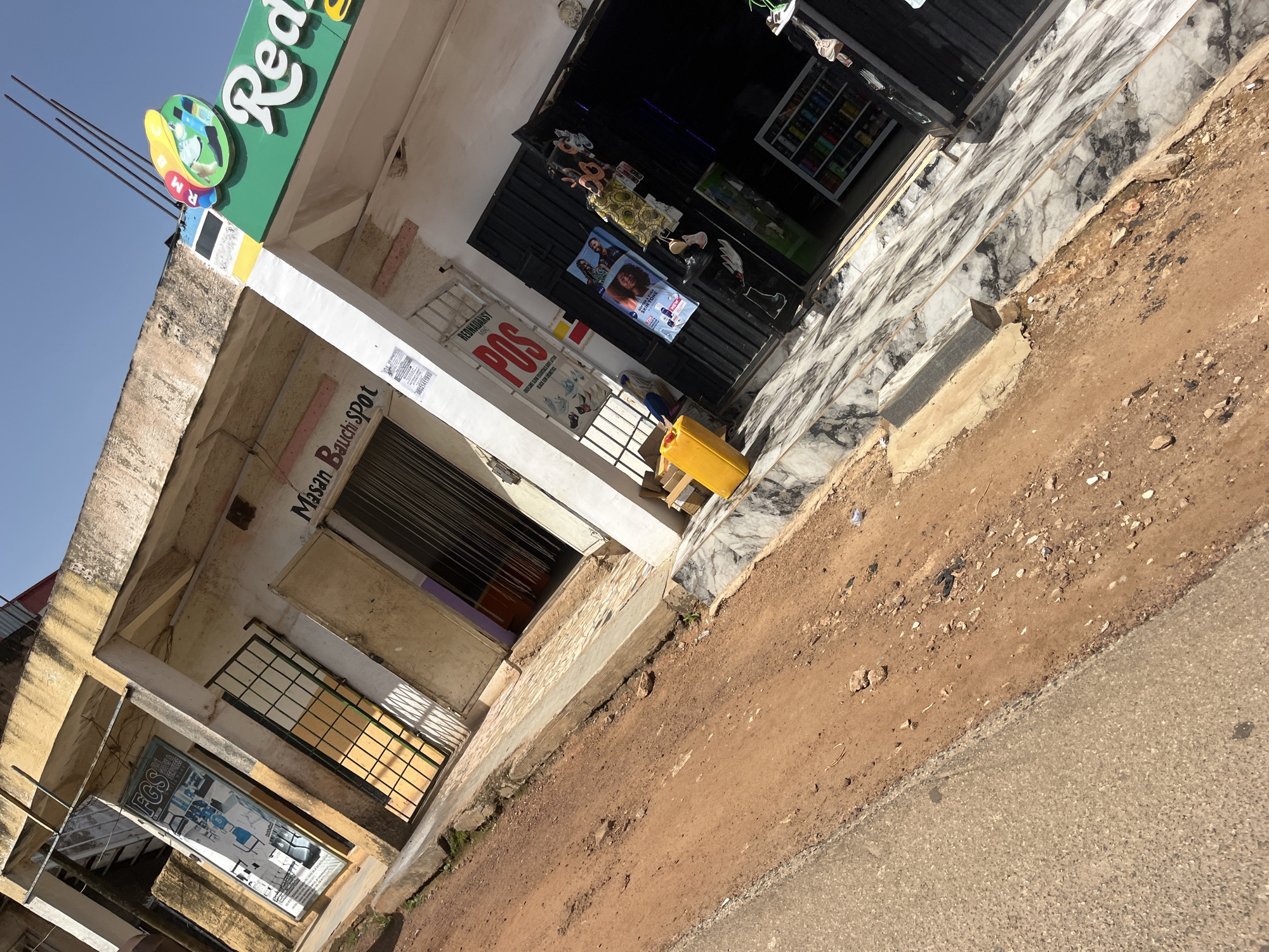 The image shows a street scene in Gura Top, Nigeria. The perspective is from a low angle, looking up towards buildings on the right side of the street. The buildings appear to be shops or small businesses.
On the far left of the image, there is a storefront with a sign that reads "FGS". Next to it is a shop with a barred window. Further to the right, another shop has a sign that says "Masan Bauchi SPOT". Adjacent to this is a shop with a green awning that has the word "Red" in white lettering, and a logo with colorful circles.
In front of the shops, there is a sidewalk paved with marble-like tiles. A yellow jerrycan is visible on the sidewalk in front of one of the shops. The street itself is unpaved and appears to be made of dirt and gravel, with some debris scattered on it. The sky is clear and blue.
No people are clearly visible in the image, but there are posters and displays in the shop windows, suggesting commercial activity. The overall impression is that of a developing urban area, with a mix of commercial establishments and unpaved infrastructure.