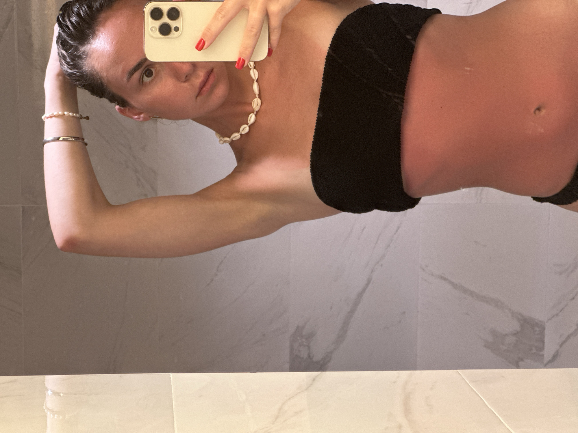 A woman is taking a selfie in a bathroom, likely indoors. She is wearing a black strapless top and a shell necklace. Her skin appears sunburnt. The background consists of light gray marble wall tiles and light beige marble floor tiles. The location is not discernible from the image.