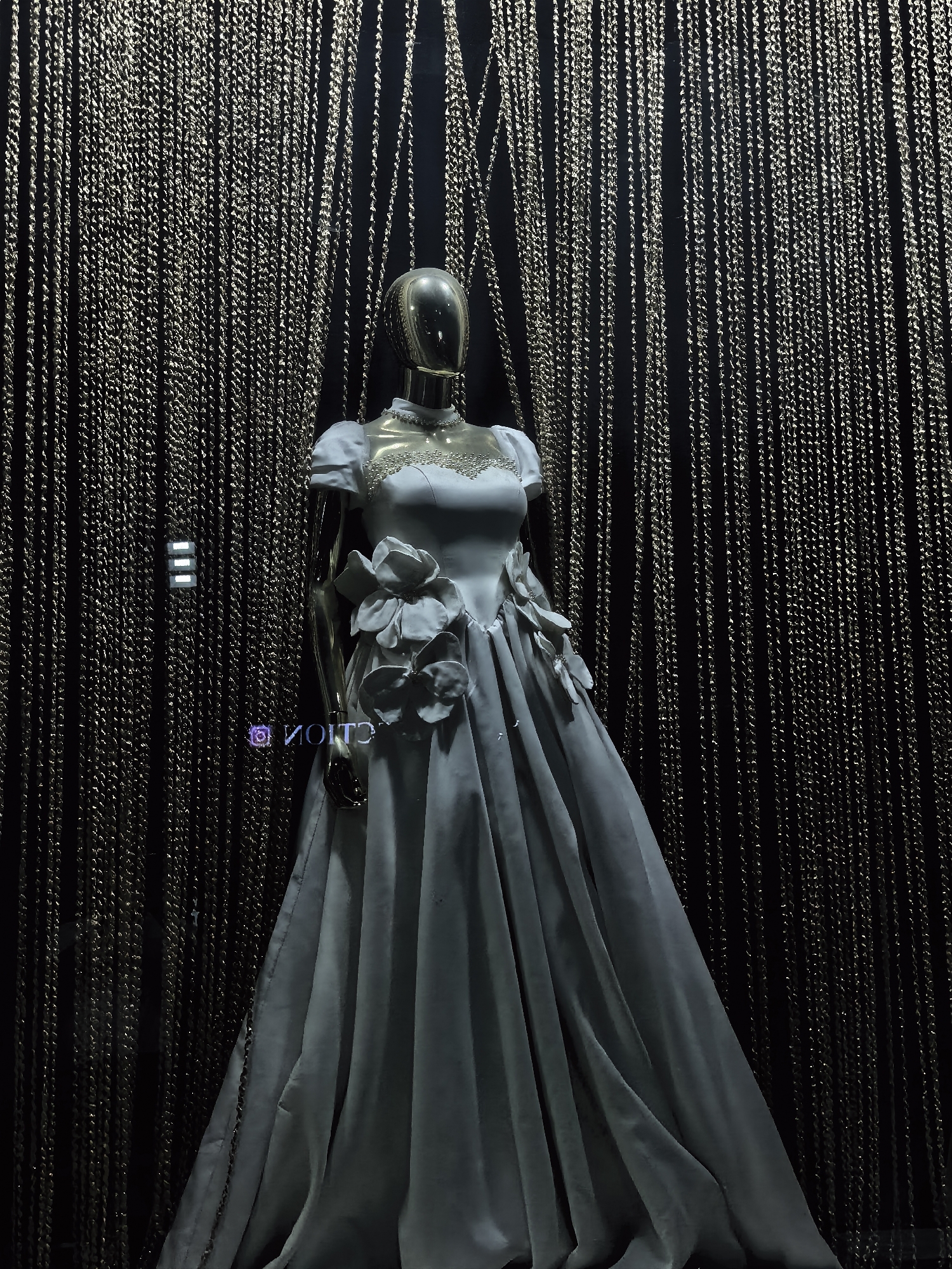 A mannequin, styled with a metallic, reflective head and a light blue, flowing gown adorned with floral embellishments, stands in a dimly lit setting. The gown features a fitted bodice with short sleeves and a voluminous skirt. The background is composed of numerous thin, hanging chains, creating a shimmering, textured curtain. To the left of the mannequin, a small, illuminated sign displays three horizontal white bars. Below this, a purple, mirrored logo reads "MOITOS". The scene appears to be a display, possibly in a shop window or exhibition. The location in Fergana City, Uzbekistan is not ascertainable from the provided image.