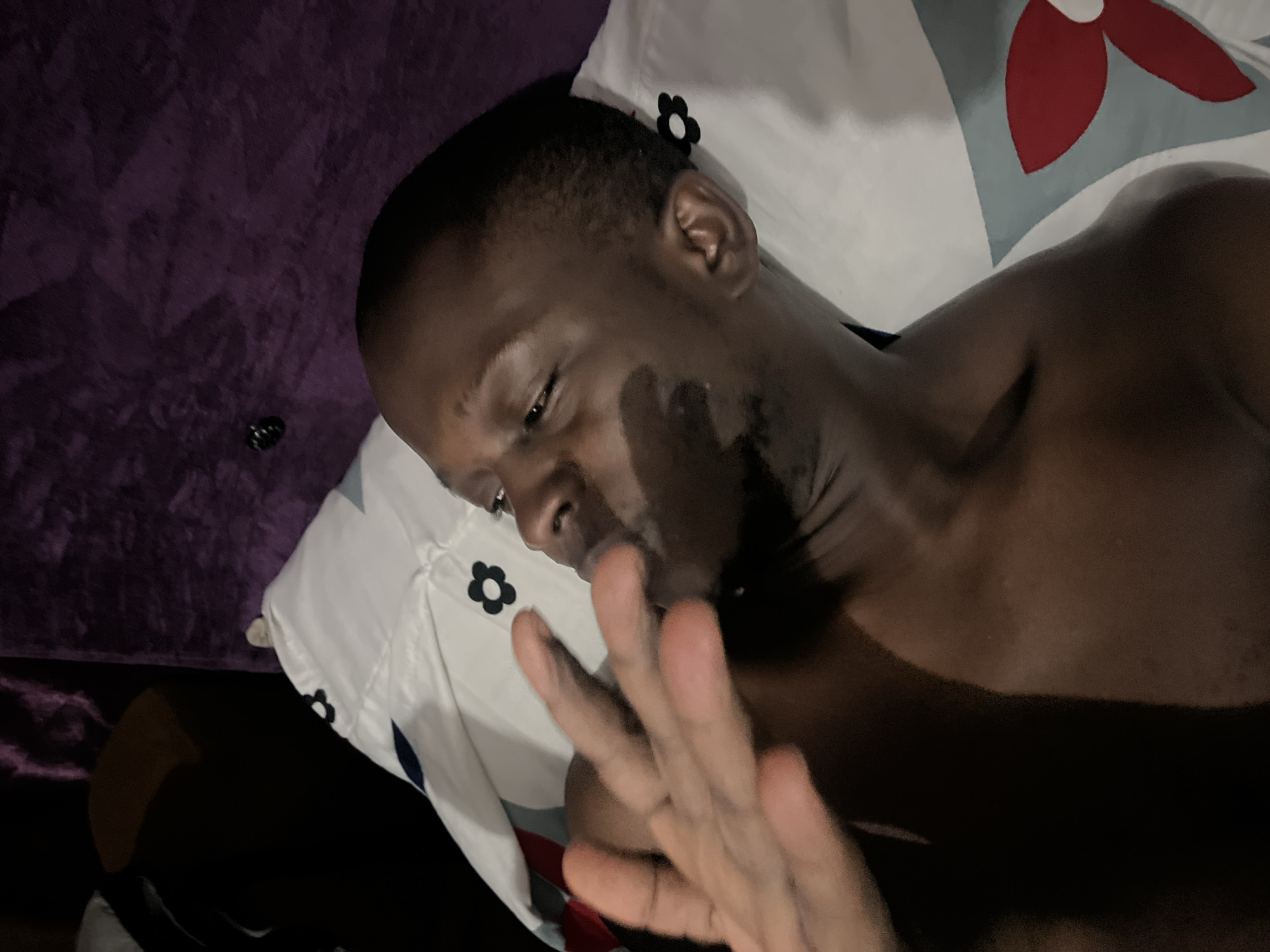 A dark-skinned man is lying on a bed with a flower-patterned pillow. He is shirtless and facing the camera. His hand is raised in front of his face. The location appears to be indoors. There is a purple wall in the background. The location in Abuja, Nigeria, is unknown.