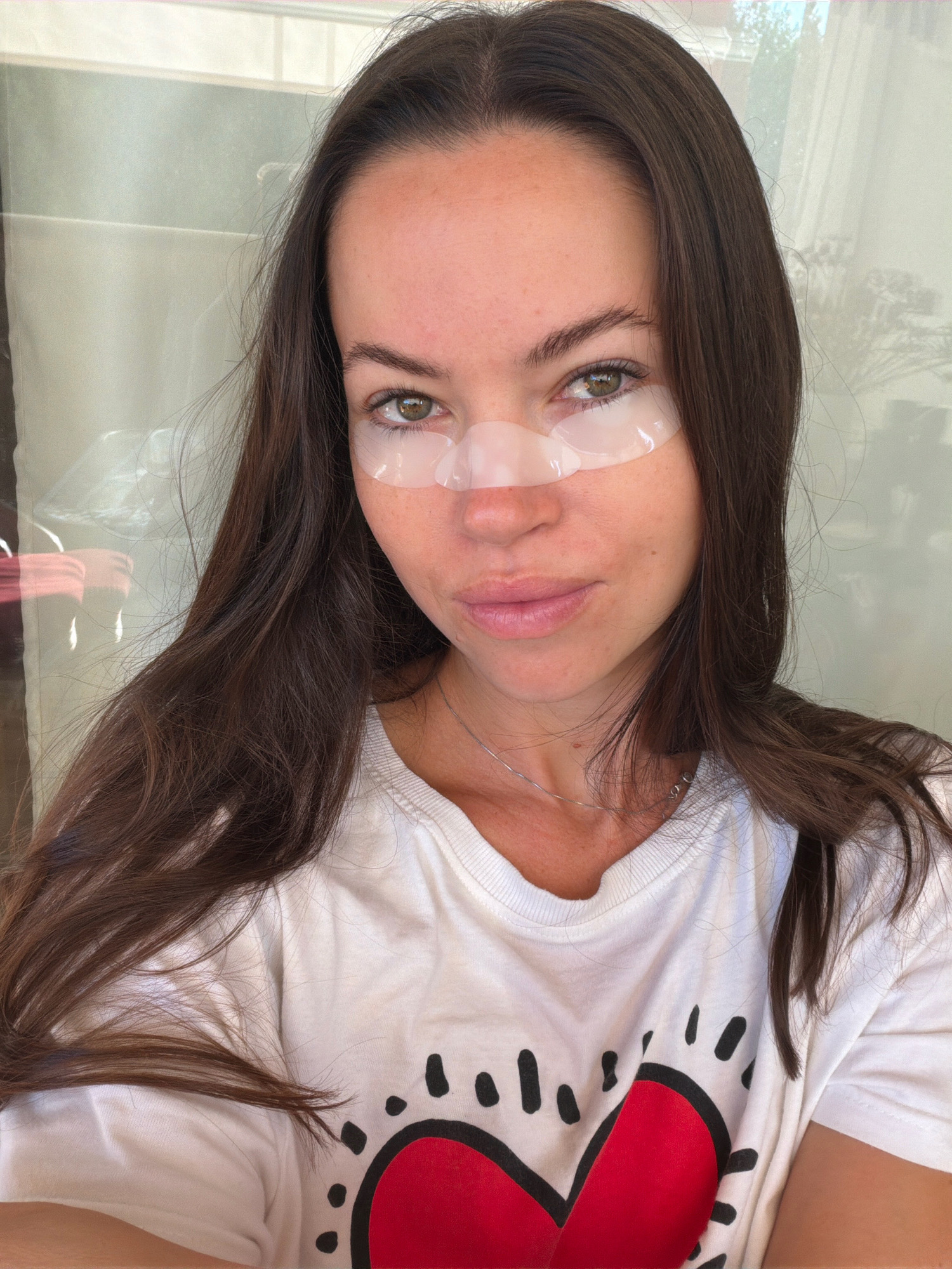 A young woman with long, dark hair and green eyes is pictured in a selfie. She is wearing a white t-shirt with a red heart graphic. Under her eyes, she has applied clear eye patches. The background appears to be an indoor setting with sheer curtains or blinds. There is no indication of the city of Salou, Spain.