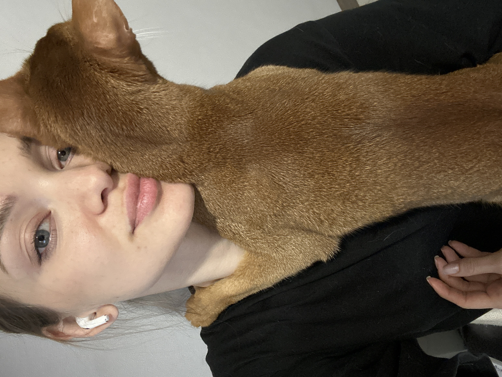 A close-up indoor photograph shows a young woman with light skin and blue eyes, wearing a black shirt and a white in-ear earbud in her left ear. An auburn-colored Abyssinian cat rests on her chest and shoulder, its head near her face. The woman's hand is partially visible in the lower right corner. The background is a plain, light-colored wall. The location in the city of Kazan, Russia cannot be derived from the image.
