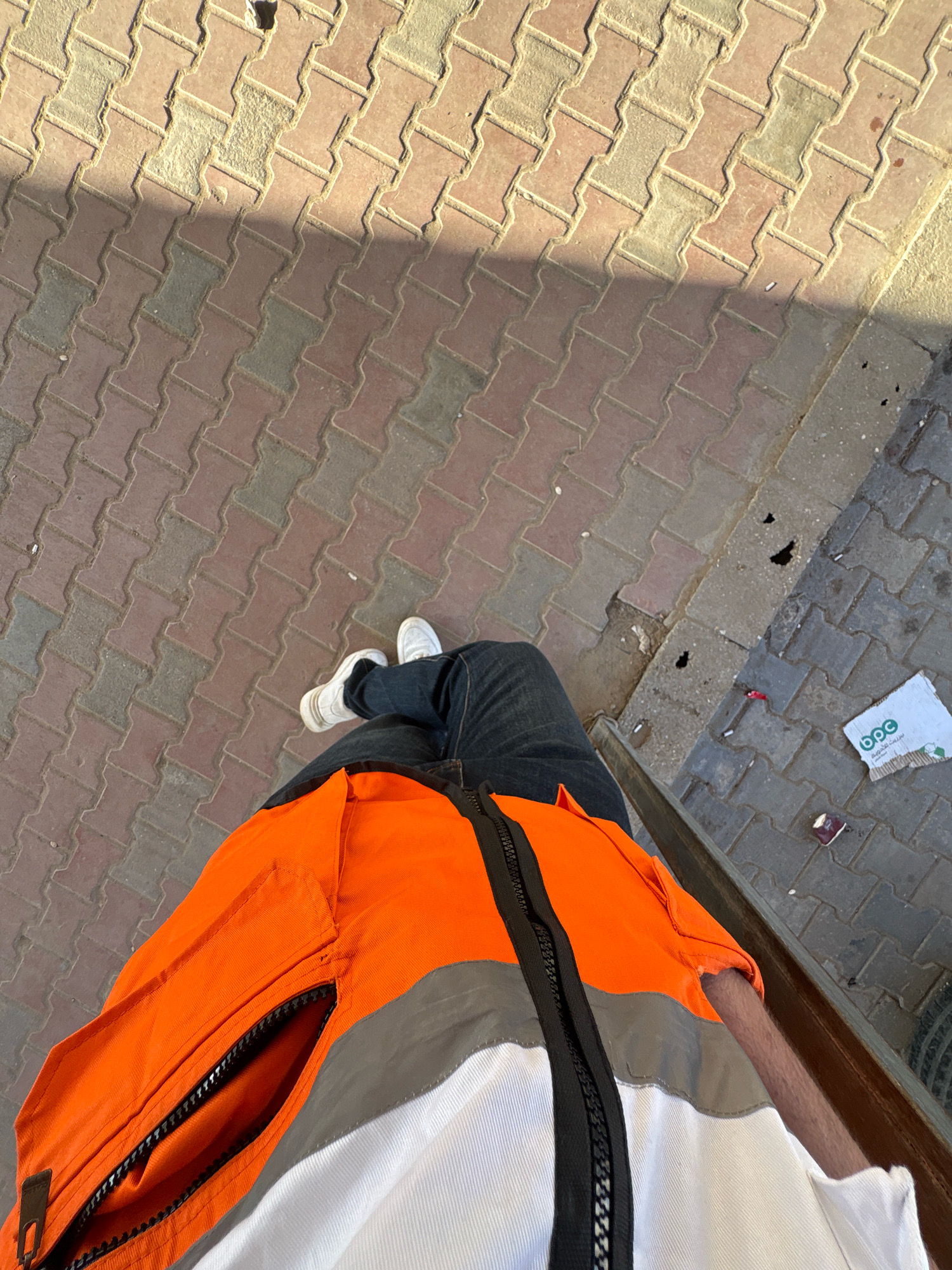 The image is a high-angle shot of a person's lower body, taken outdoors. The person is wearing dark jeans, white sneakers, and a high-visibility jacket with orange, gray, and white sections. The jacket has a black zipper. The background is a street paved with interlocking brick pavers, some of which appear worn or damaged. The pavers are arranged in a herringbone pattern. A shadow is cast diagonally across the pavers from the upper left to the lower right of the frame. In the lower right portion of the image, there are scattered pieces of debris on the pavement, including a crumpled white piece of paper with a green logo that appears to say "b.o.c.", cigarette butts, and a small red object. The overall environment suggests an urban or semi-urban setting, possibly a sidewalk or a street in the city of Gaza, Palestinian Territories, given the context. The lighting suggests it is daytime, likely with sunlight creating the shadow.
