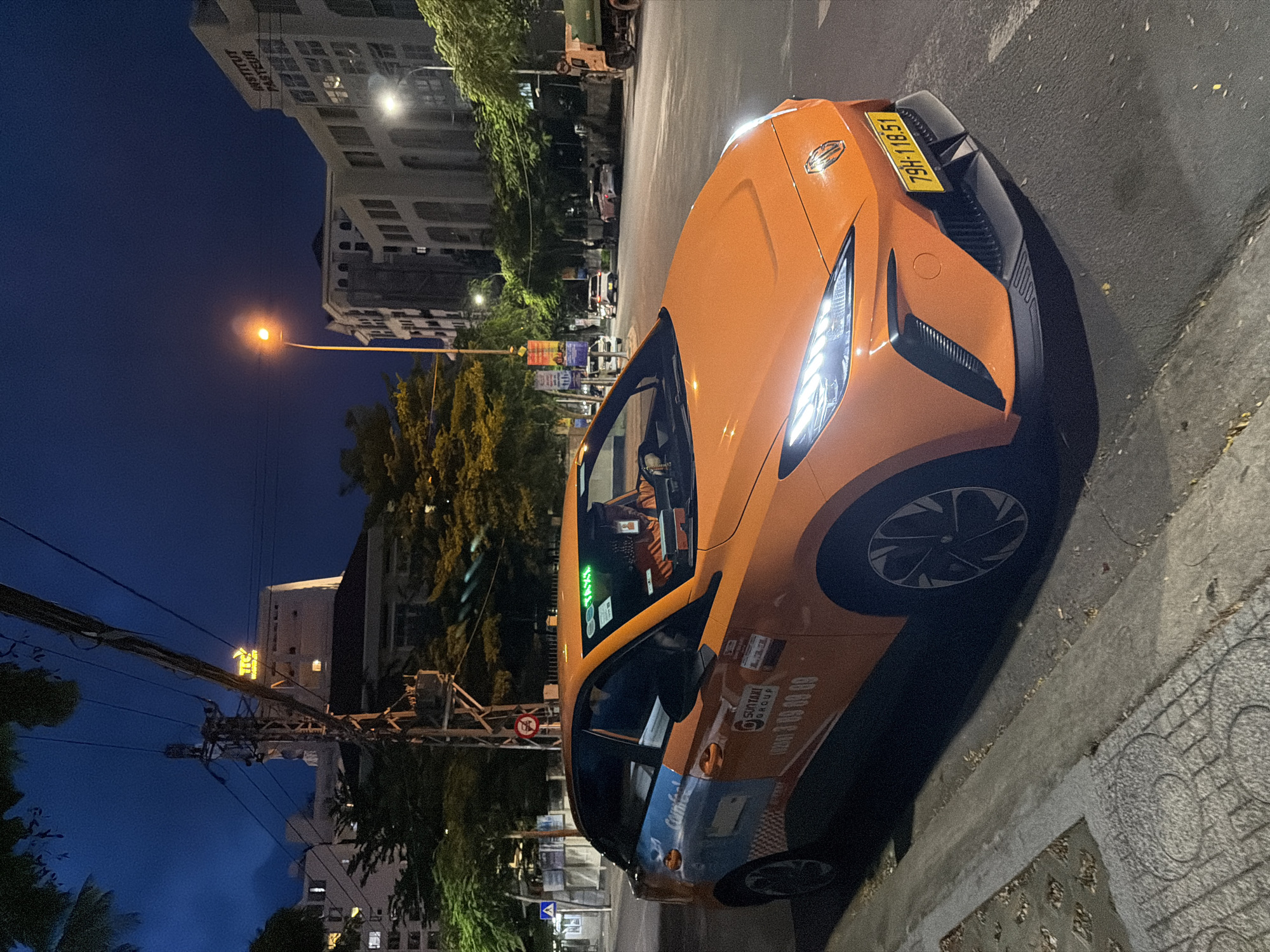 A bright orange electric taxi is parked on the side of a street at night in Nha Trang Ward, Vietnam. The taxi has a blue and white stripe on its side and a phone number printed on the door. The license plate of the taxi reads "79H-118.51". In the background, there are buildings, trees, streetlights, and a utility pole. The sky is dark blue, indicating nighttime. The taxi is angled towards the camera, with its headlights illuminated.