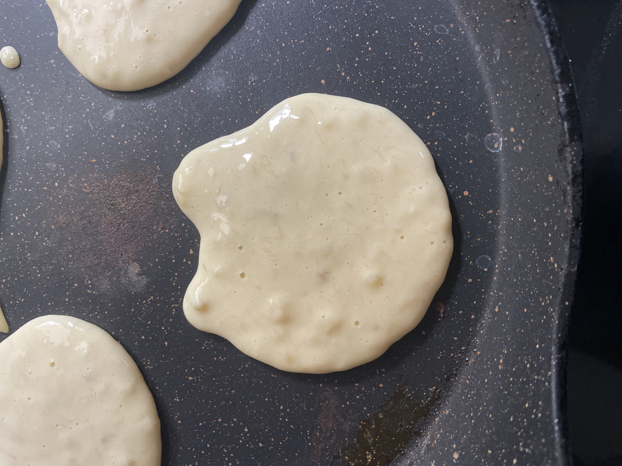 The image shows several uncooked pancakes on a dark-colored, speckled frying pan. The pancakes are light in color with a slightly uneven surface. There are no people visible in the image. The background is out of focus and does not provide any clues about the location. It is not possible to determine if the image was taken in Denpasar, Indonesia.