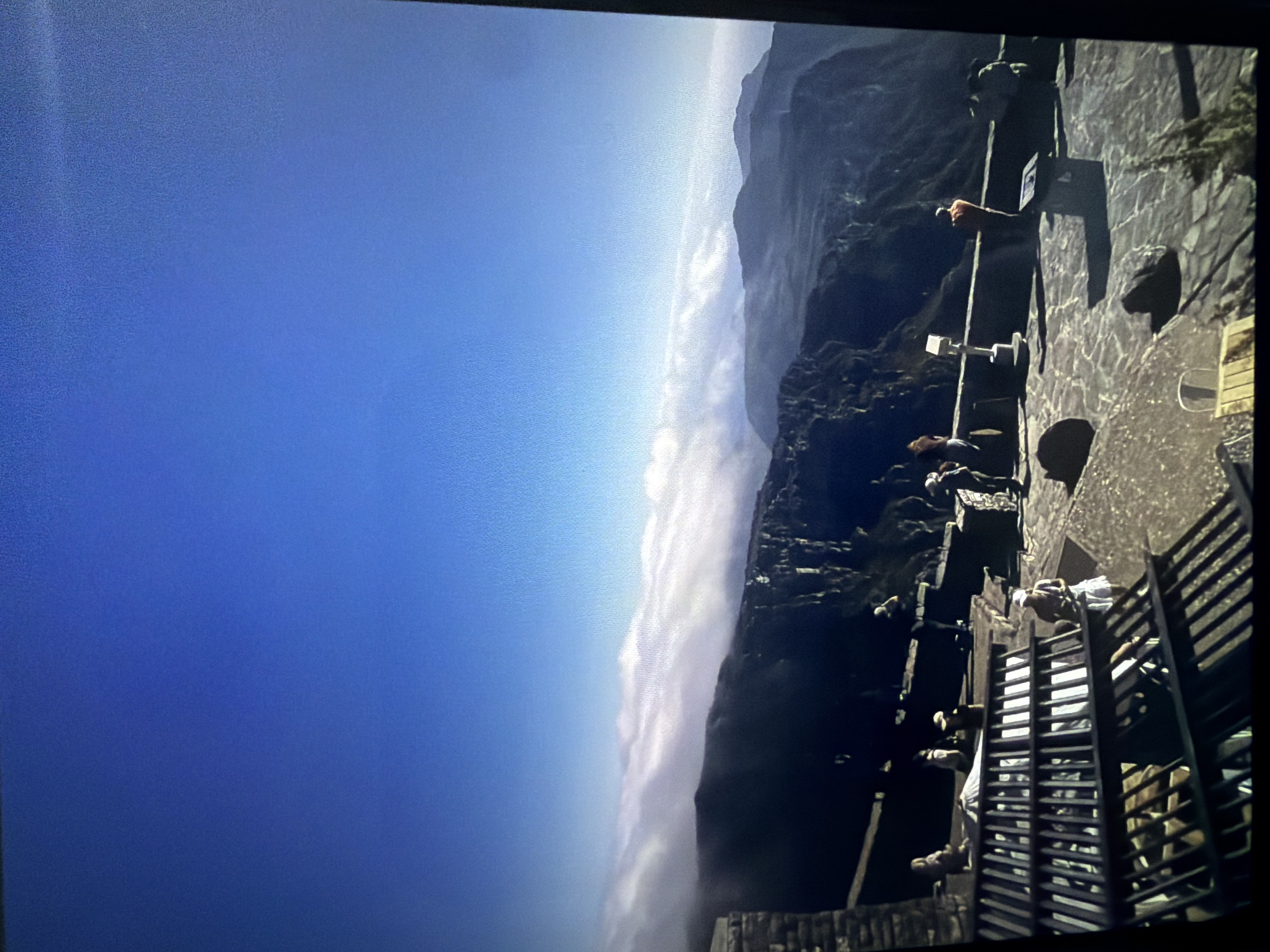 The image, when correctly oriented, presents a majestic high-altitude view from what appears to be a mountain summit or an observation deck, likely in Cape Town, South Africa, consistent with a vantage point like Table Mountain.
In the foreground, a robust stone wall or ancient-looking structure forms a barrier, equipped with dark railings. Scattered along this structure are several people. One individual, wearing a bright red or orange top, is prominently seen leaning on a railing, gazing out at the scenery. Another person dressed in white is positioned near a dark, ladder-like structure that seems to be part of the wall or a descent path. Other less distinct figures can be seen observing the view.
The scene dramatically opens to a vast, clear blue sky dominating the upper portion of the frame. Below the sky, a thick, undulating layer of white clouds or mist stretches horizontally, creating a stunning cloud inversion effect, with the viewpoint appearing to be above the clouds. Dark, rugged mountain peaks and ridges emerge from and rise above this cloud layer on the right side, adding to the dramatic landscape. The lighting suggests it is daytime, possibly morning or late afternoon, with soft illumination. The activity centers around sightseeing and enjoying the panoramic vista. There is no visible text in the image.