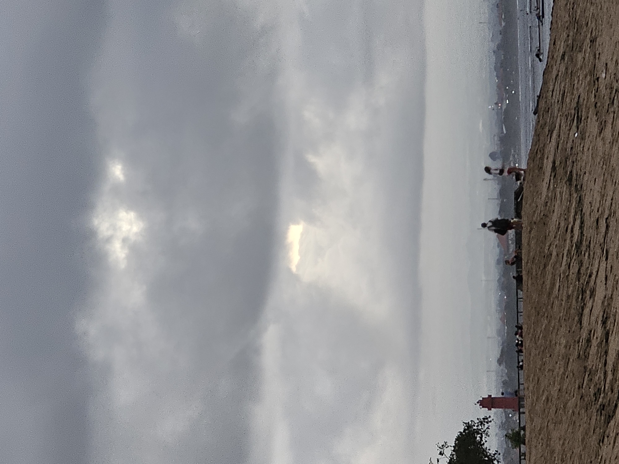 The image depicts a hazy, overcast sky over a coastline in Denpasar, Indonesia. In the foreground, a sandy embankment slopes down towards a body of water. Along the embankment, several people are visible, some standing and some sitting, observing the scene. To the left, a red lighthouse stands prominently. In the middle ground, the water is calm, and several boats are moored or sailing in the distance. The background features a hazy cityscape with distant buildings and structures, suggesting an urban or suburban area. The overall atmosphere is subdued due to the cloudy weather.