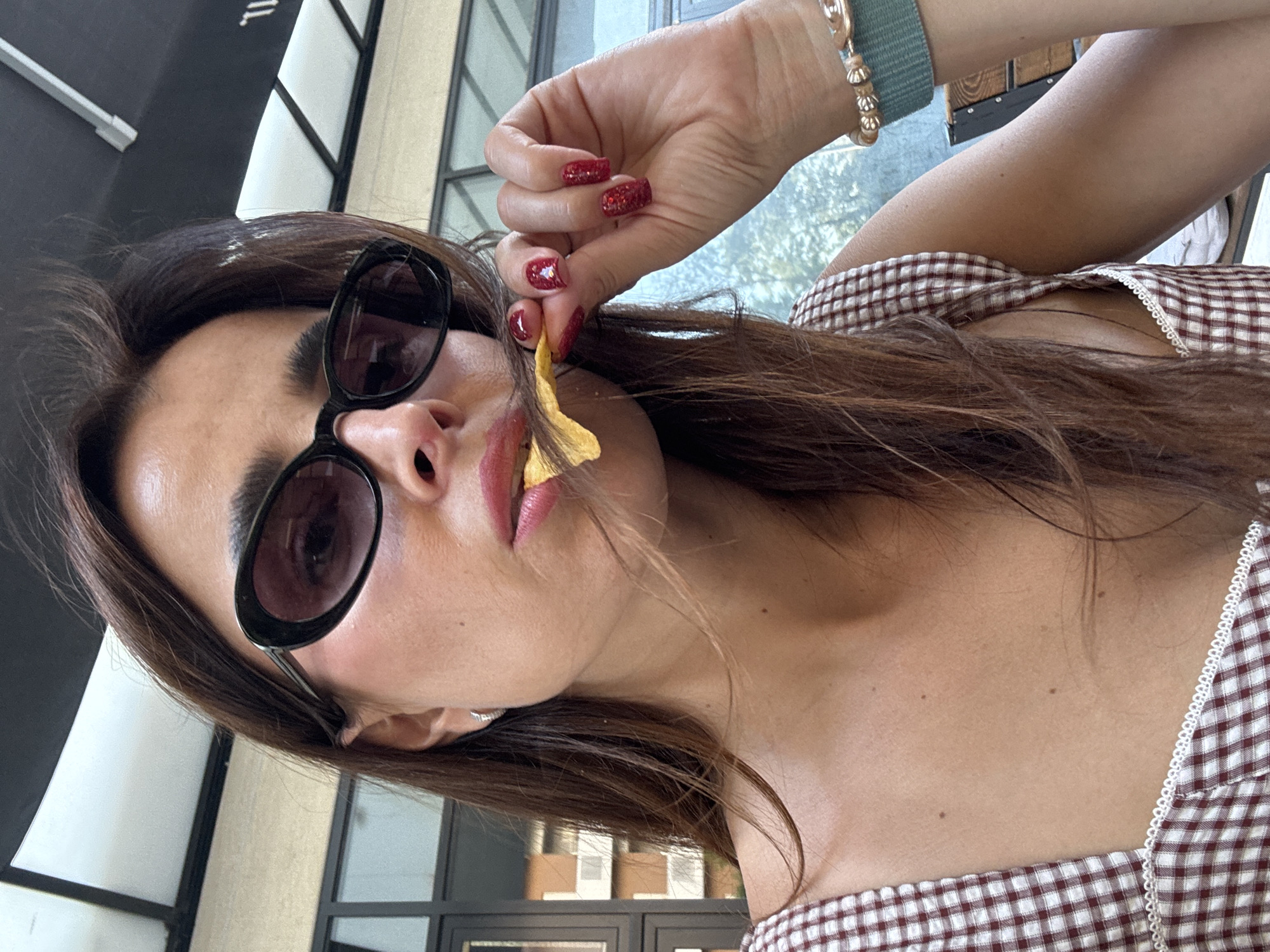 The image shows a close-up of a woman's face as she eats a chip. She is wearing sunglasses, has long brown hair, and is dressed in a plaid top. The background appears to be an outdoor seating area, possibly a cafe or restaurant, with some buildings visible in the distance. The image does not contain any specific visual cues that would allow for the identification of its location within Podgorica, Montenegro.