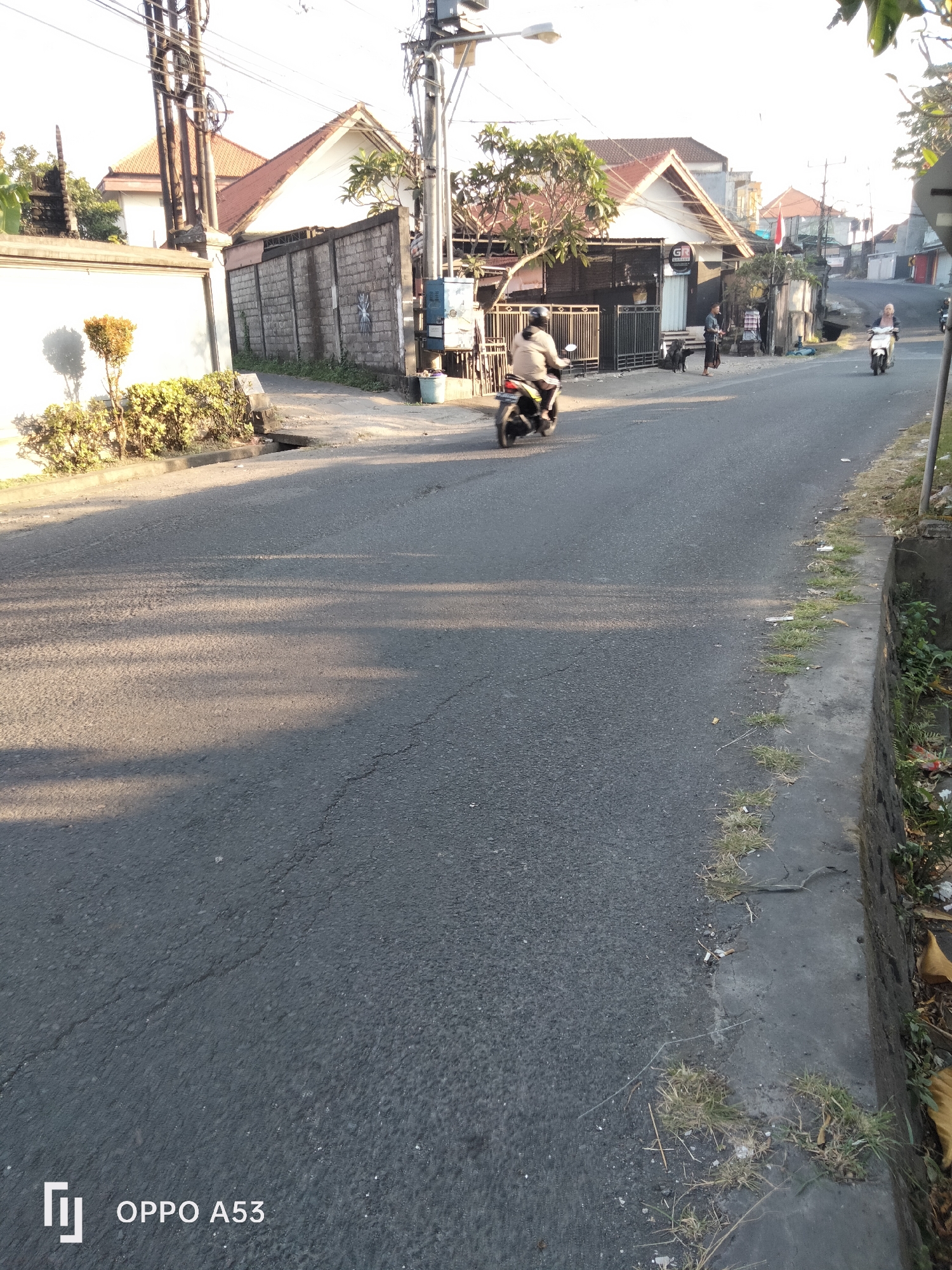 The image depicts a street scene in Denpasar, Indonesia, with a focus on the road and its surroundings. In the foreground, the asphalt road is cracked in several places and shows evidence of wear and tear. Sunlight casts dappled shadows across the road from trees and overhead wires. On the right side of the road, a concrete curb is visible, with patches of grass growing through cracks and along the edges. Debris, including cigarette butts and dried leaves, is scattered on the curb and the adjacent ground.
Further down the road, several individuals are present. On the left side of the road, a person is riding a motorcycle, moving away from the camera. The motorcycle appears to be dark in color, and the rider is wearing a light-colored jacket and helmet. On the right side of the road, another person is riding a scooter, also moving away from the camera. This individual is wearing a light-colored top and dark pants. In the background, on the left side of the road, a building with a white facade and a red-tiled roof is visible, bordered by a grey stone wall and a white wall. There are also utility poles with numerous wires crisscrossing overhead. On the right side of the road, further in the distance, more buildings and structures are visible, suggesting a residential or commercial area. A small trash can is also present near the utility pole. The overall impression is that of a typical urban street on a sunny day in Denpasar.