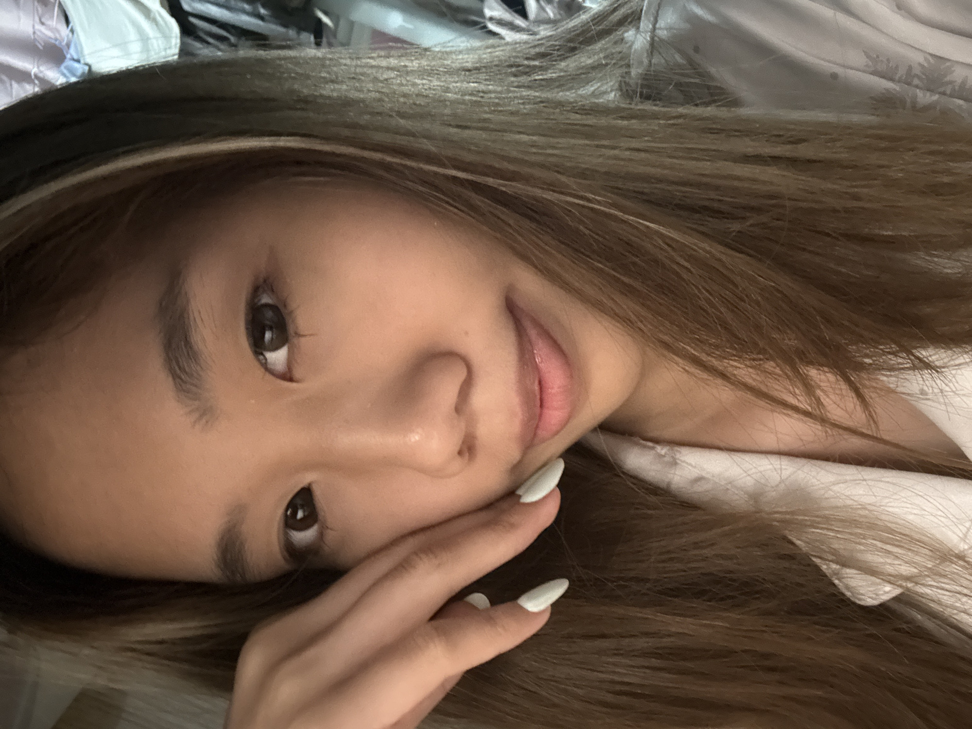 A close-up, rotated shot of a young Asian woman with long, brown hair featuring blonde highlights. She is leaning her head on her hand, which has long, white, almond-shaped nails. The background is out of focus and appears to be clothing, with a white garment visible near her shoulder. The image doesn't contain enough information to confidently assert any location in the city of Nan'an District, China.
