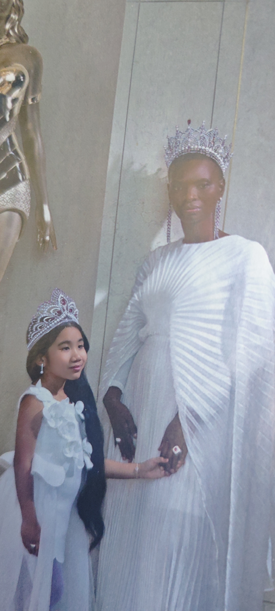 Two figures, a child and an adult, both wearing crowns and light-colored dresses, stand holding hands in front of a light-colored wall. To the left of the pair is a partially visible golden mannequin. The location is unknown; there is no visible indication that this image was taken in Novosibirsk, Russia.