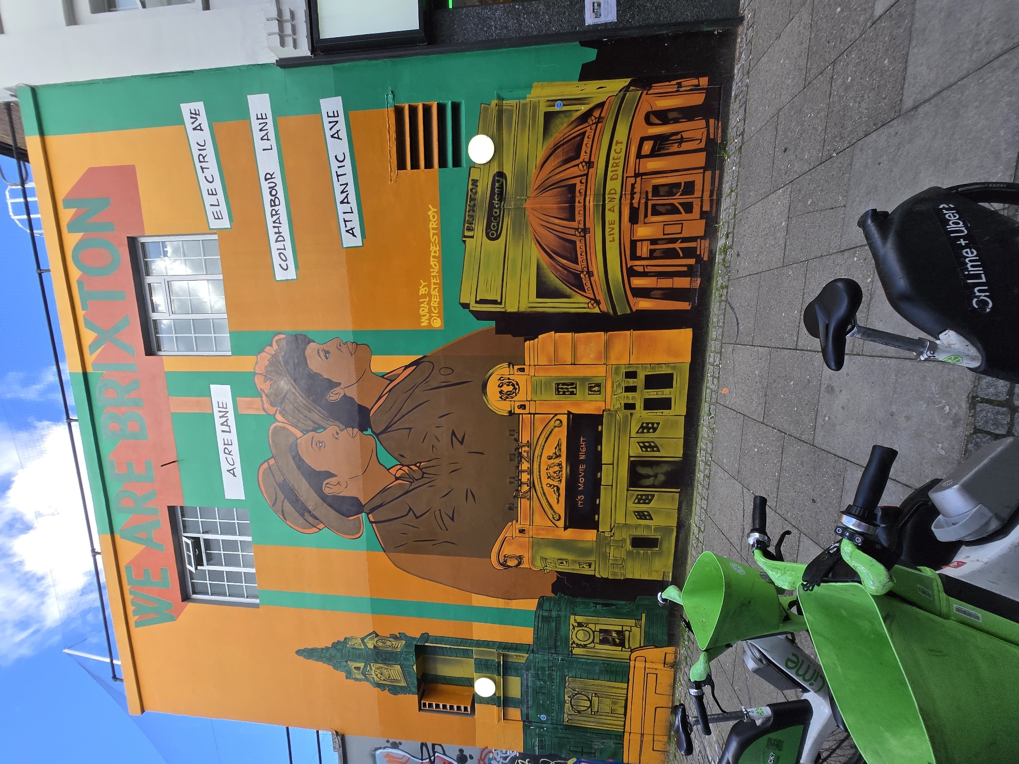 This image depicts a vibrant mural covering the facade of a building in Brixton, London, United Kingdom. The mural features prominent text stating "WE ARE BRIXTON" at the top in green letters against an orange background. Below this, four white signs with black text indicate local street names: "ACRE LANE," "ELECTRIC AVE," "COLDHARBOUR LANE," and "ATLANTIC AVE."
The mural's central section includes two stylized profile busts of individuals, a man and a woman, depicted in brown tones with hats, facing right. To the left, a green and yellow clock tower structure is painted. In the mid-right, a yellow and orange building resembling the Ritzy Cinema is shown with the text "RITZY" and "IT'S MOVIE NIGHT." Further right, another yellow and orange building with a distinctive domed roof represents the Brixton Academy, labeled "BRIXTON o'academy" and "LIVE AND DIRECT." The mural also incorporates two real windows into the building's upper section; one is partially open. A smaller inscription on the right indicates "MURAL BY @ICREATENOTDESTROY." Two white circular light fixtures are integrated into the mural on the building wall.
In the foreground, several green and black Lime rental bicycles are parked on a grey tiled pavement. One bike clearly displays "On Lime + Uber?" on its rear fender. The sky above the building is blue with scattered white clouds. A portion of an adjacent light-colored building is visible to the right, and some graffiti is present on a wall to the left of the main mural. The scene is illuminated by bright daylight.