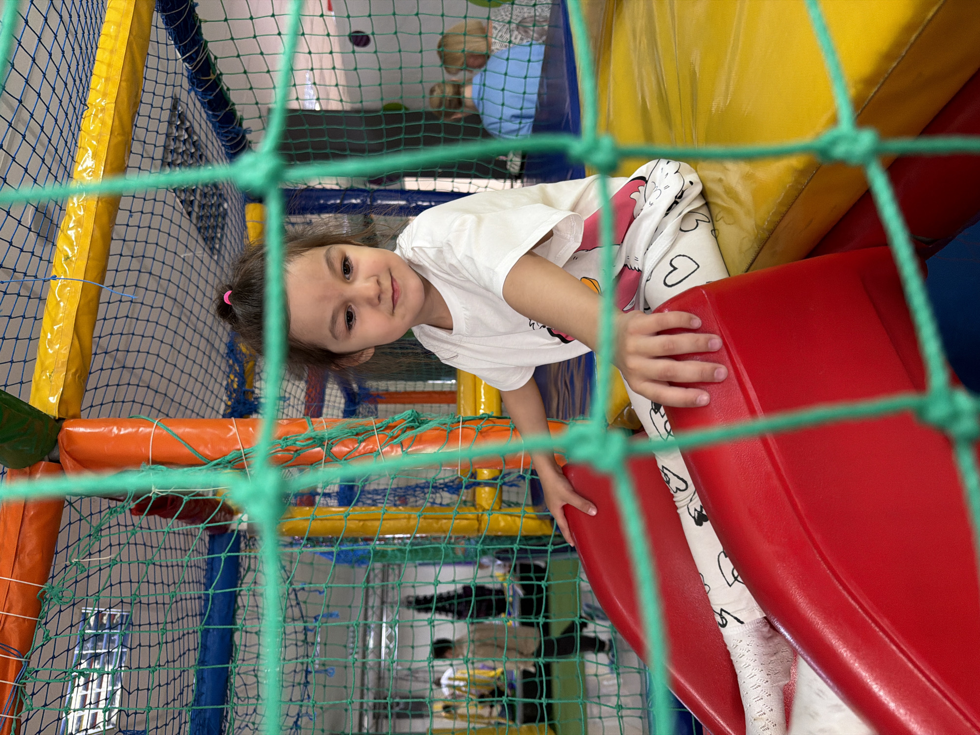 A child is seen through green netting, wearing a white shirt and patterned pants, standing on a red and yellow play structure. The netting suggests an enclosed play area, possibly a playground or indoor play center. Other colorful padding and play structures are visible in the background. The location is Chaikovsky, Russia.