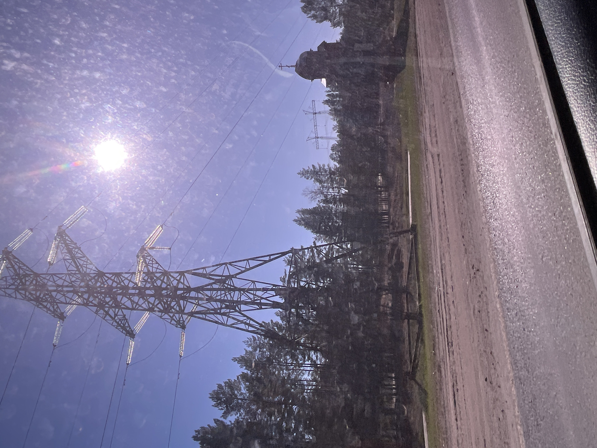 View through a dirty window of a rural landscape in Noviy, Russia. Bright sun in the upper left corner of the frame. A high-voltage power line tower is visible on the left of the frame, with transmission lines stretching towards the upper right corner. A forest extends along the right side of the image with what appears to be an orthodox church partially visible in the trees. A road or dirt path runs alongside the forest. The location in the city of Noviy, Russia can not be confirmed with this image.
