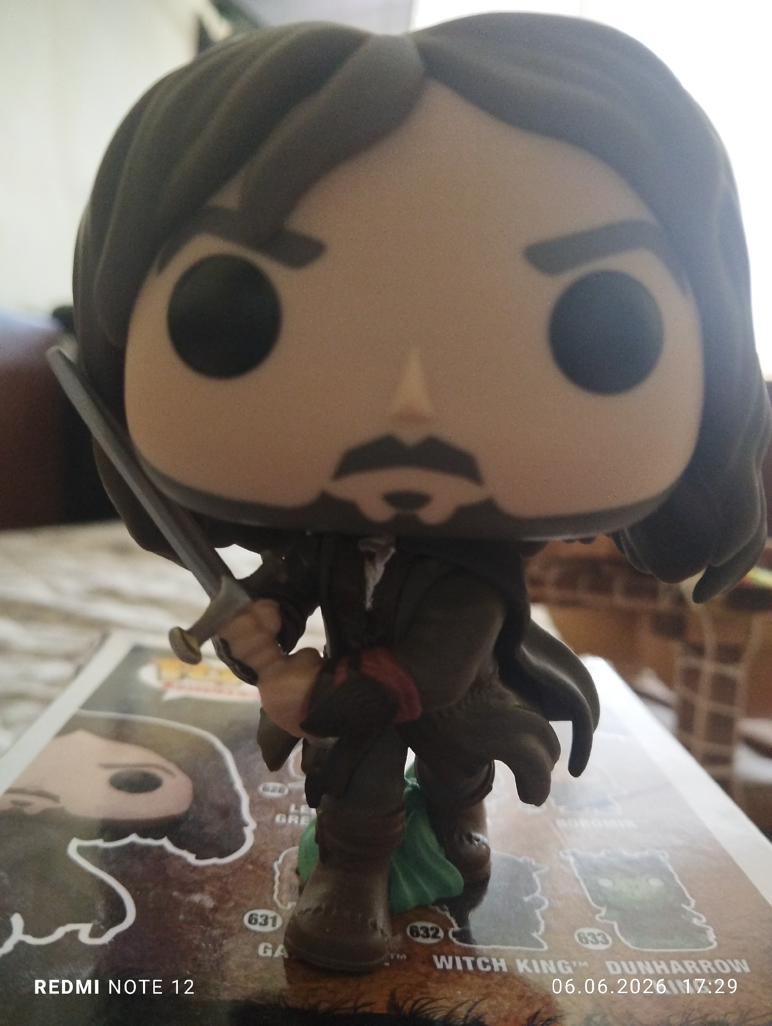 A Funko Pop vinyl figure, identified as Aragorn from The Lord of the Rings, is centrally featured, depicted standing upright and holding a silver sword with a gold hilt. The figure possesses dark brown hair, a tan skin tone, black dot eyes, and a black printed beard and mustache. It is attired in a dark brown outfit with a reddish detail on the right arm, and a green fabric detail is visible near its feet. The figure stands on what appears to be a packaging insert or a display card, which includes outlines of other Funko Pop characters and numerical identifiers such as '626', '631', '632', and '633'. Visible text on this surface includes 'WITCH KING™ DUNHARROW', 'BOROMIR' (partially obscured), and an illustration of another Funko Pop figure on the left. The background is blurred, indicating an indoor environment with indistinct shapes and varying light levels. The image bears two watermarks: 'REDMI NOTE 12' in the bottom left corner and '06.06.2026 17:29' in the bottom right corner. The scene is set in an indoor location in Burgas, Bulgaria.