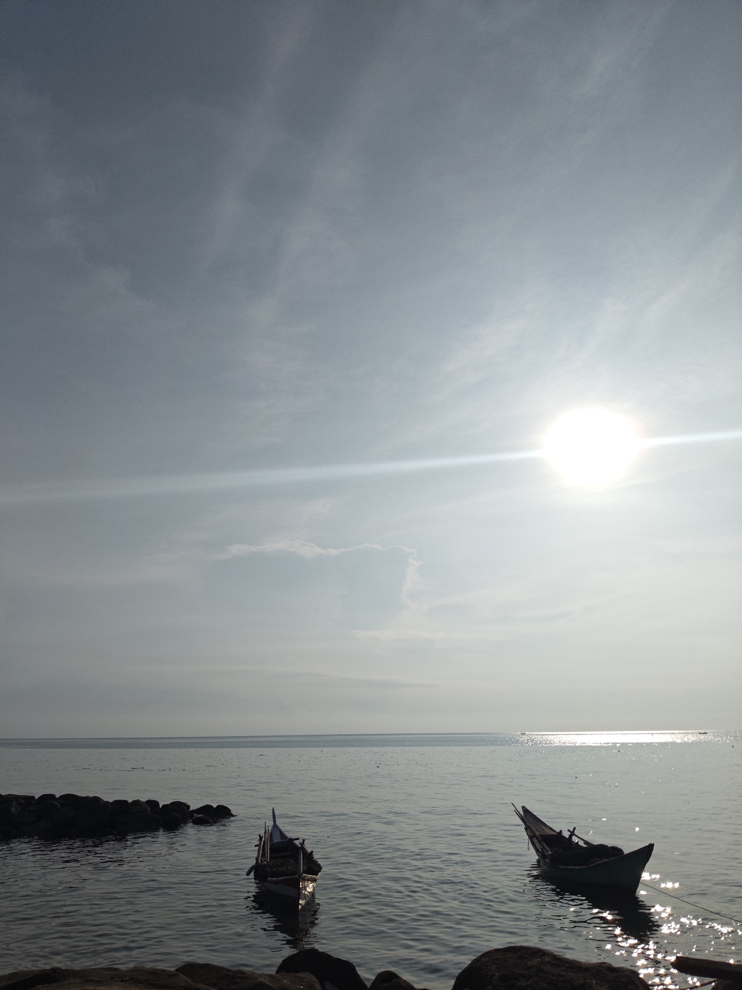 The photograph depicts a coastal scene in Lhokseumawe, Indonesia, under a bright, hazy sun. The sky is predominantly light blue with thin cloud cover and a visible sun flare. The sea is calm with reflective glints of sunlight, indicating a mild ripple on the surface. Two small fishing boats are anchored in the water, both appearing unmanned. In the foreground, there are rocks at the water's edge. The horizon is visible but hazy, suggesting distance. The lighting suggests it is daytime, possibly midday. The composition focuses on the serene, natural environment with human presence manifested only through the boats.
