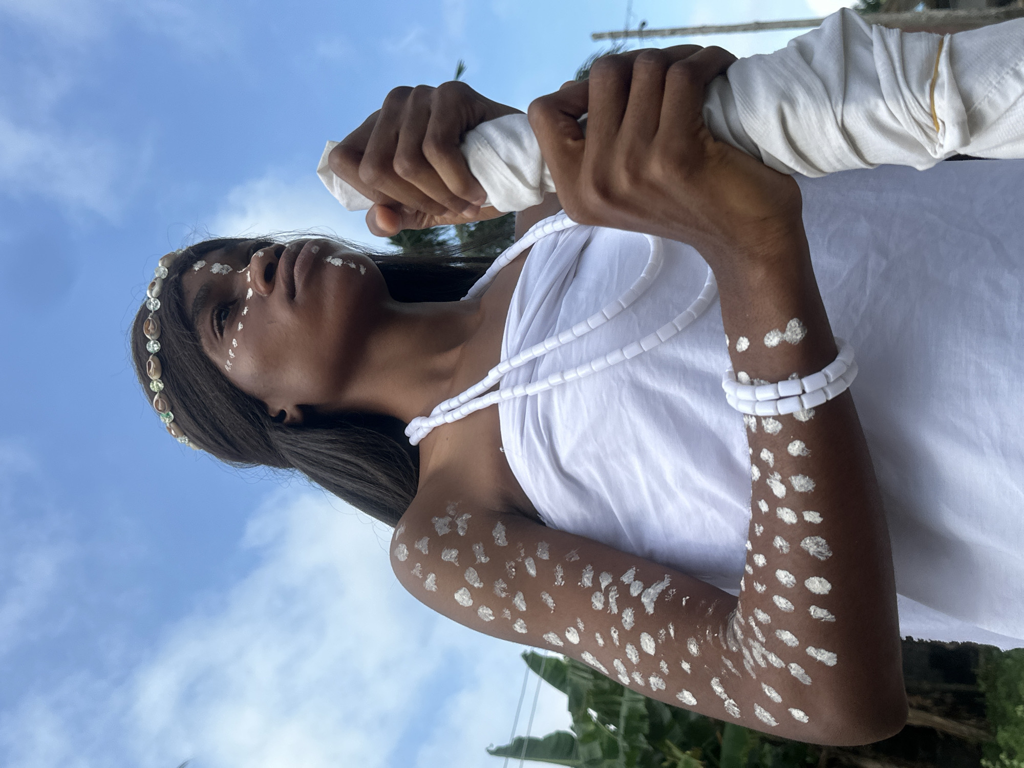 In Badagry, Nigeria, a young woman with dark skin stands outdoors, looking to her right. She is adorned with white face paint in a geometric pattern, with dots on her cheeks and a line on her forehead and chin. She wears a white headpiece adorned with shells and a white beaded necklace. Her left arm is covered in white paint dots, and she has a white beaded bracelet on her left wrist. She is wrapped in a white cloth and holds a white fabric object in her right hand. The background consists of a blue sky with scattered clouds, and blurred greenery is visible to the left.