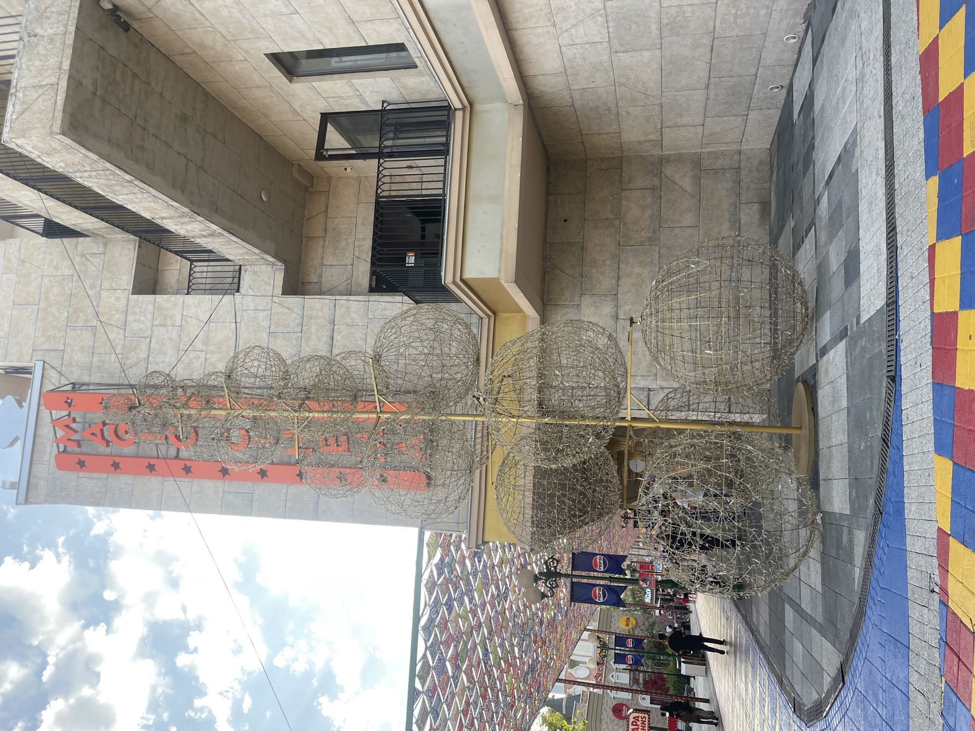 The image depicts a street scene in Tashkent, Uzbekistan, with a focus on a building facade and an outdoor art installation. The building, constructed with light-colored, textured stone blocks, features multiple stories with balconies. One prominent balcony is visible, with a black metal railing and a concrete floor. A sign with red lettering and star shapes is mounted vertically on the building, reading "MAGIC."
In the foreground, a large, wireframe sculpture resembling a stylized tree adorned with illuminated spheres is the central art piece. The spheres are constructed from a light-colored wire mesh, possibly containing embedded lights. The sculpture is gold-colored at its base and trunk.
The street in front of the building is paved with a pattern of gray, black, and white square tiles, bordered by strips of blue and yellow tiles, creating a vibrant, checkerboard-like effect. Along the street, shops and banners are visible, including one with the "Pepsi" logo. Several individuals are present on the street, some walking, others standing. Overhead, a colorful canopy of patterned fabric stretches across the street, adding to the lively atmosphere. The sky above is partly cloudy.