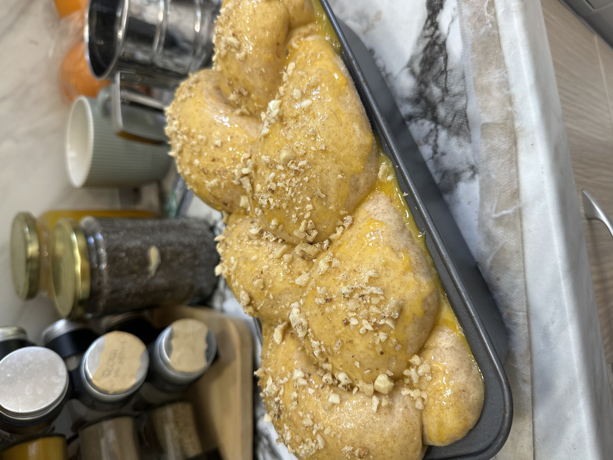 A close-up shot captures a loaf of challah bread in a dark metal baking pan. The bread has a golden-brown, egg-washed sheen and is sprinkled with chopped nuts. The pan sits on a countertop with a faux marble pattern. In the background, there are various kitchen items including spice jars, a glass jar filled with honey and other kitchen appliances and utensils. The image does not provide any information about the location in Varna, Bulgaria. There are no people visible in the image.