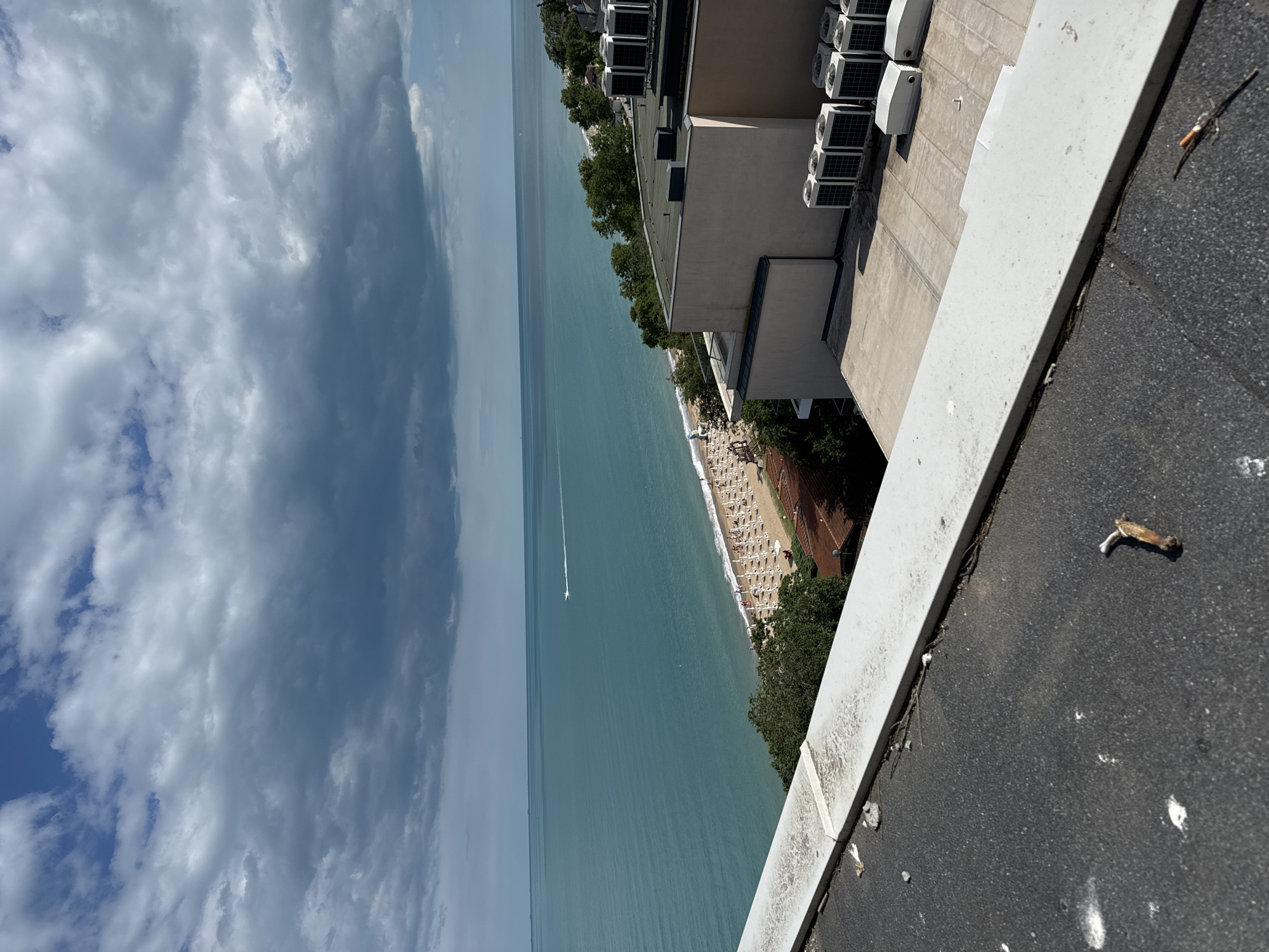 The image captures a high-angle view of a coastline, likely in Varna, Bulgaria. The scene is dominated by the sea, a beach, and rooftops. The sea is a vibrant turquoise and stretches to the horizon, meeting a dramatic sky filled with dark, heavy clouds interspersed with patches of blue. A small white boat is visible on the water, leaving a faint wake.
The beach is lined with rows of white umbrellas and lounge chairs, suggesting it's a popular recreational area. To the right of the beach, a densely wooded area separates it from buildings. In the foreground, the edge of a dark, asphalt-like rooftop is visible, with a white parapet. Beyond this, the flat roofs of several buildings are apparent, equipped with numerous outdoor air conditioning units. Some of these units are arranged in rows, while others are housed in white rectangular enclosures. The architecture appears modern. The overall impression is one of a coastal resort area under a moody sky.