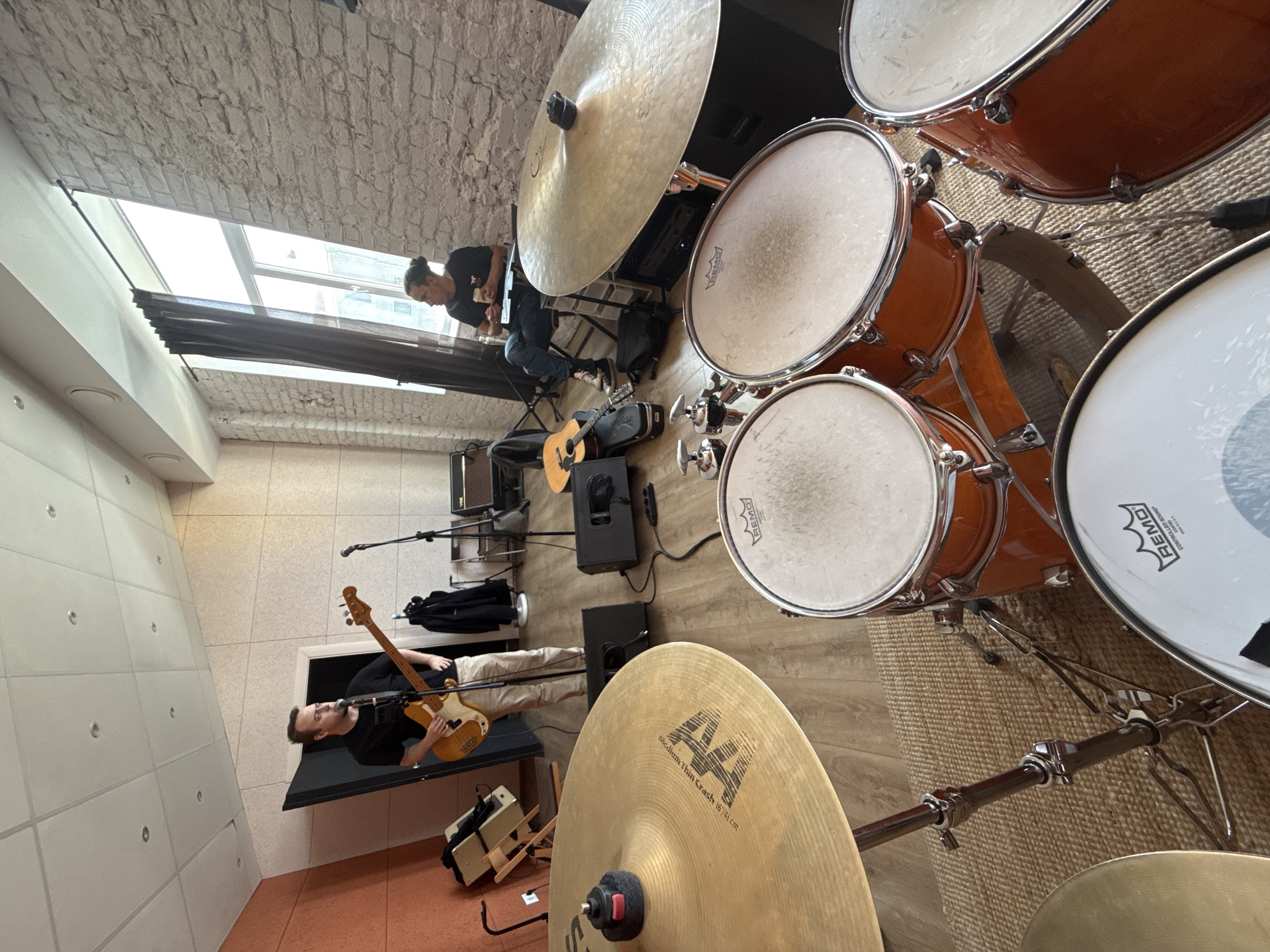 A drum kit dominates the right side of the image. Adjacent to the kit, a man in a black shirt and light-colored pants plays a bass guitar near a small black table. Further back, another man in a black shirt and jeans sits and appears to be working on a laptop or tablet. An acoustic guitar sits on the floor near the seated man. Musical equipment, including amps and speakers, are scattered around the floor. The floor is light-colored wood. The room has a high ceiling with a textured, potentially brick surface. A window with blinds is visible in the background. This scene appears to be taking place in a rehearsal room. While the location cannot be definitively determined to be Moscow, Russia, based on the image alone, the possibility cannot be ruled out.