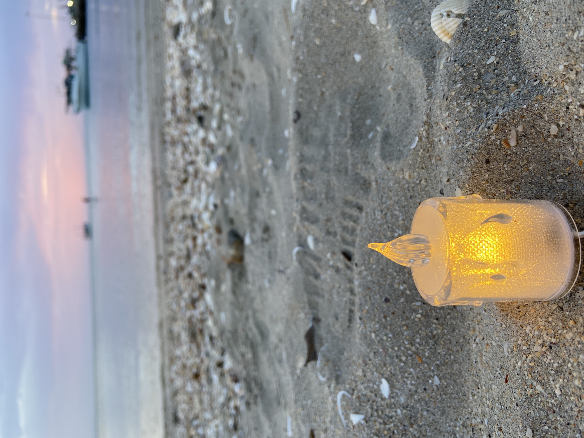 A close-up, low-angle shot captures a small, battery-operated LED candle resting on a sandy beach in Juan Griego, Venezuela. The candle, made of translucent plastic with a flickering yellow LED light, is positioned in the foreground. Its sides are textured, resembling drips of wax. The sand around the candle is a mixture of fine grains and small shell fragments. In the mid-ground, the beach continues, with more shells scattered across its surface. The background features the calm, reflective water of the sea, with a soft, pastel-colored sky in shades of pink and purple, indicative of either dawn or dusk. A few blurred boats are visible on the horizon. The overall impression is one of peaceful serenity.