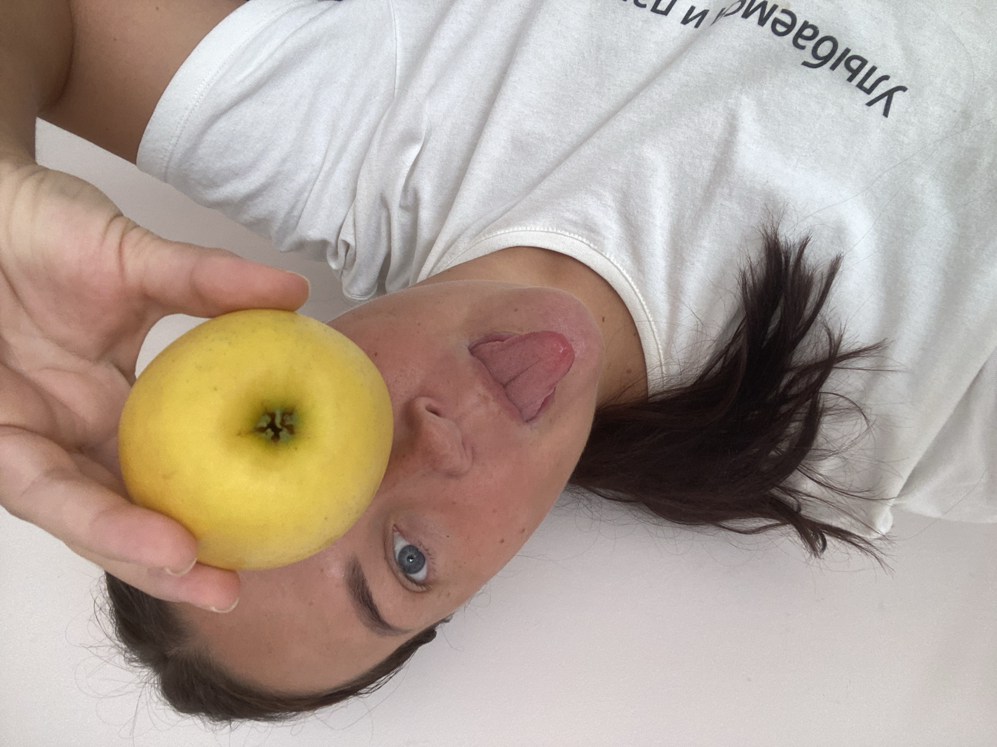The image shows a young woman holding a yellow apple in front of her eye and sticking her tongue out. She is wearing a white t-shirt with Russian text on it. The background is a plain white wall. There is no indication of the city of Sochi, Russia in the image.