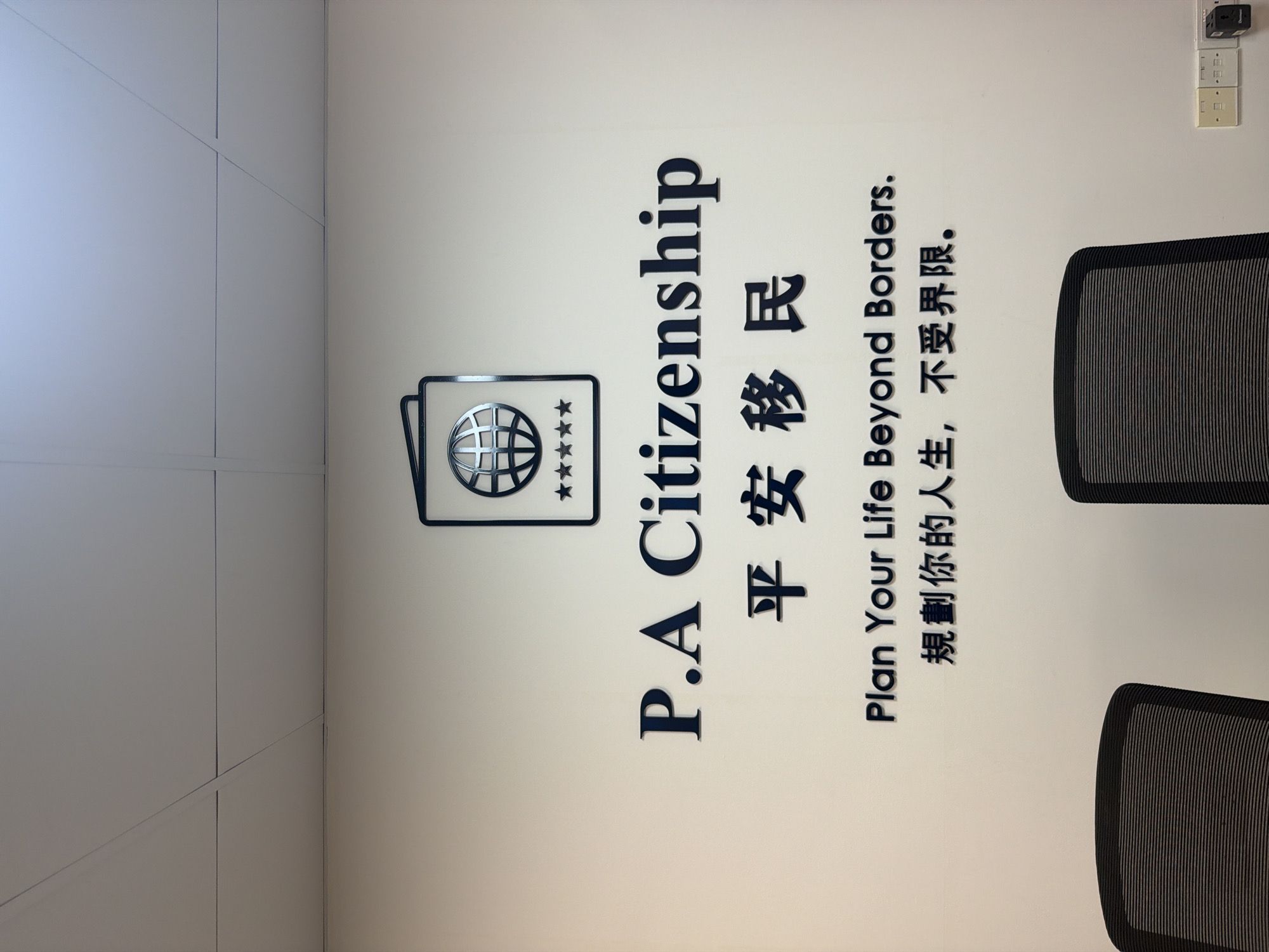 A close-up, vertically-oriented indoor shot features a wall with text and a passport graphic. The wall is light-colored, possibly white, and appears to be part of an office space, suggested by two black office chairs visible on the right. The text, rendered in a dark color like navy blue, reads "P.A Citizenship" in English, with the Chinese characters "平安移民" below. Further text reads "Plan Your Life Beyond Borders." followed by "規劃你的人生,不受界限." in Chinese. To the left of the English and Chinese text, a passport-style graphic is present, featuring a globe and five stars. The upper right corner shows electrical outlets. The location is speculated to be in Business Bay, United Arab Emirates.
