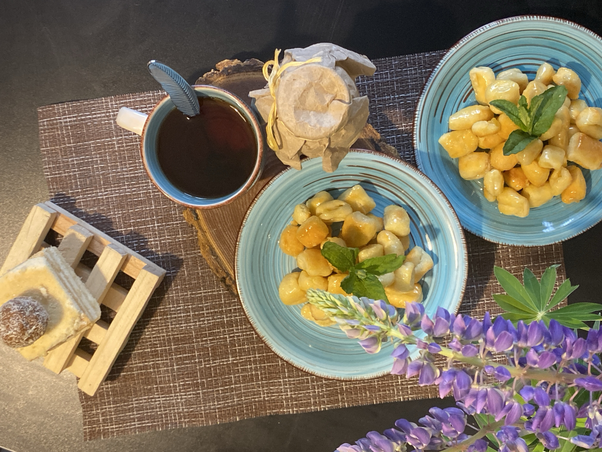 The image shows a table setting with various food items and a cup of tea. Two light-blue plates are filled with fried dough pieces, garnished with mint leaves. A paper-wrapped container, possibly holding honey or jam, sits next to a light-blue mug filled with tea and a spoon with a feather-shaped handle. A small wooden stand holds layered pastries topped with a ball. A sprig of purple lupine flowers lies near one of the plates. The setting is on a woven placemat on a dark surface, suggesting a meal or snack in the city of Марковский, Russia.