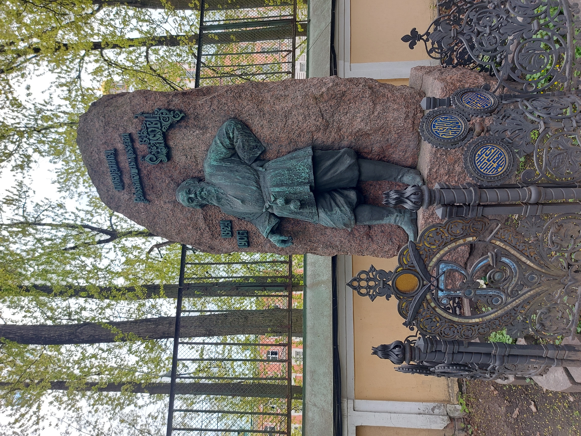 The image depicts the tomb of Vasily Yakovlevich Jacobi, located in the Necropolis of Masters of Arts at the Tikhvin Cemetery in Saint Petersburg, Russia. A bronze figure of Jacobi is sculpted on a large boulder. Text is inscribed on the boulder above the figure. The dates 1834 and 1906 are inscribed below the figure. The tomb is surrounded by an ornate wrought-iron fence. Trees and a building are visible in the background.