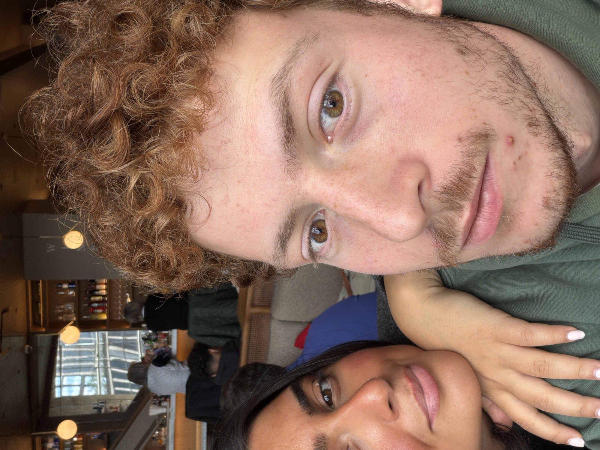 A selfie-style photograph depicts two individuals in an indoor setting. The individual on top has curly reddish-brown hair, fair skin, and light stubble. The person below has dark hair and tan skin. Their hand is placed between the two subjects. A portion of an indoor space is visible in the background, suggesting a restaurant or bar-like setting with multiple people and a well-lit shelf containing bottles. The location of the photo is not discernible in the image.